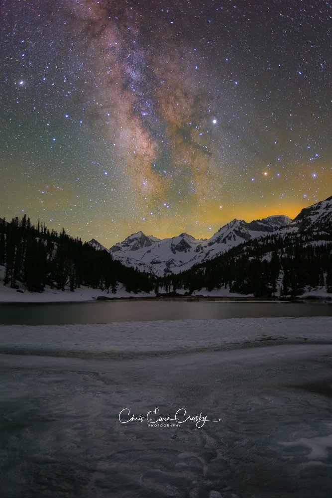 Vertical night photo of the Milky Way over a frozen alpine lake and snow-capped Sierra mountains.