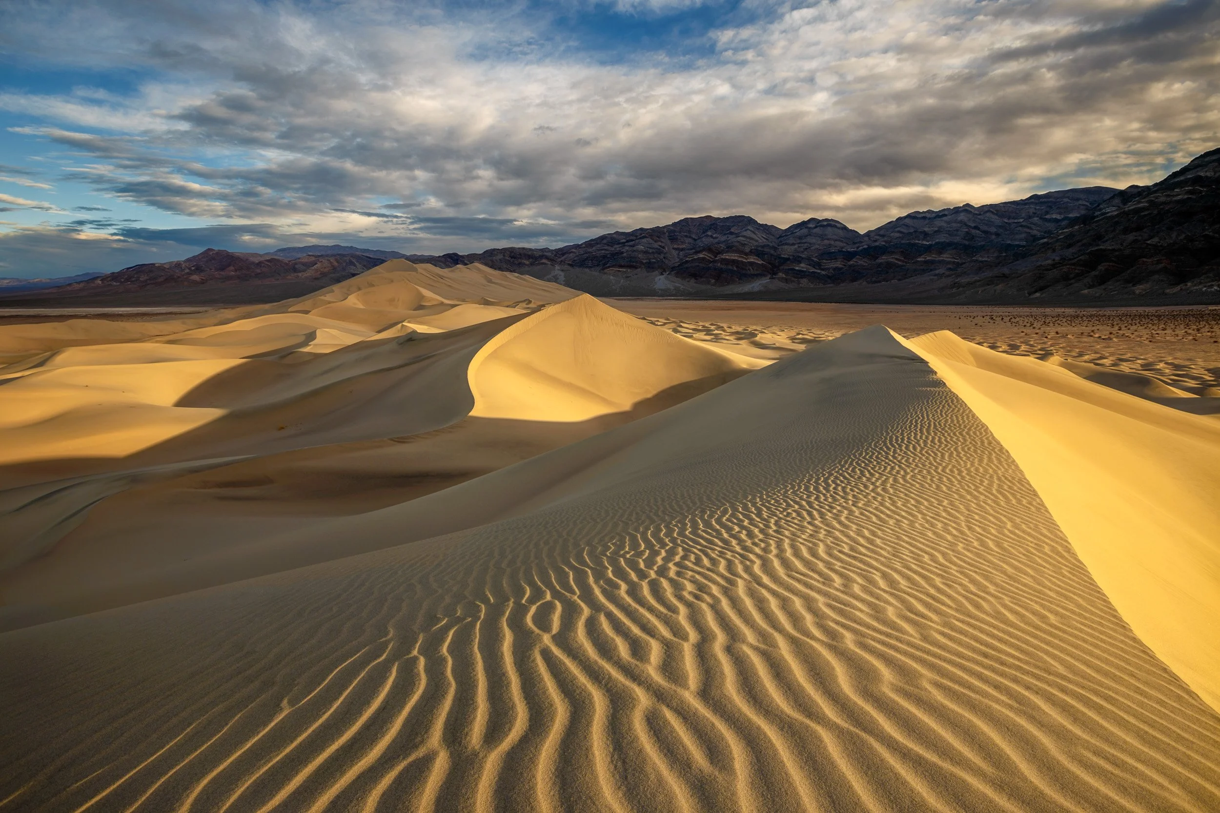 Horizontal landscape photograph of golden sunlight highlighting delicate sand ripples on the ridge of Eureka Sand Dunes with mountain ranges under a cloudy sky in Death Valley National Park by Chris Ewen Crosby.
