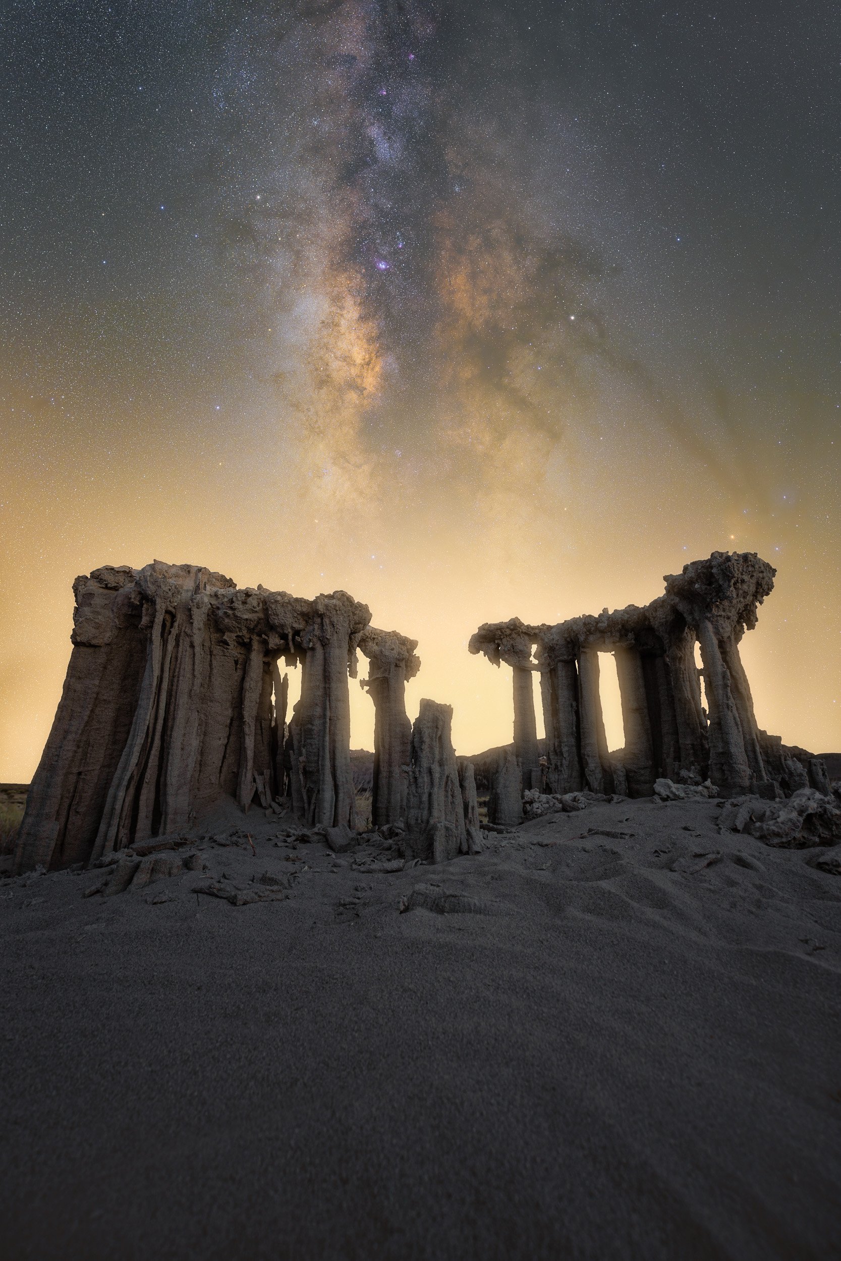 Vertical night astrophotography photograph of ancient sand tufa columns silhouetted against a glowing horizon and the Milky Way galaxy at Mono Lake in the Eastern Sierra by Chris Ewen Crosby.