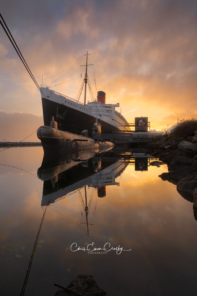 A horizontal photography print of the Queen Mary ocean liner at sunrise, reflected in the calm waters of the Long Beach harbor with a soft orange and blue sky.