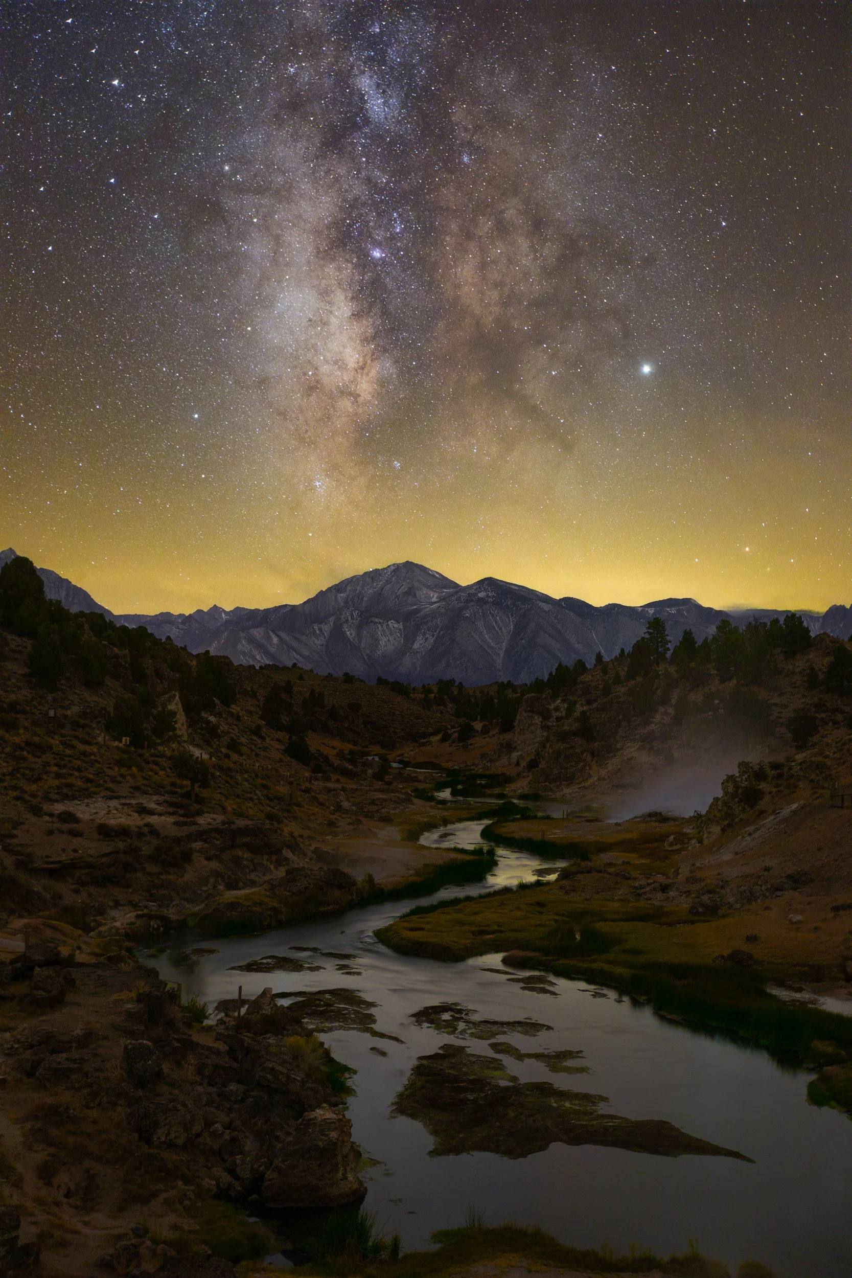 Vertical night astrophotography photograph of winding geothermal creek waters leading toward Eastern Sierra mountains beneath the Milky Way core at Hot Creek Geological Site near Mammoth Lakes by Chris Ewen Crosby.