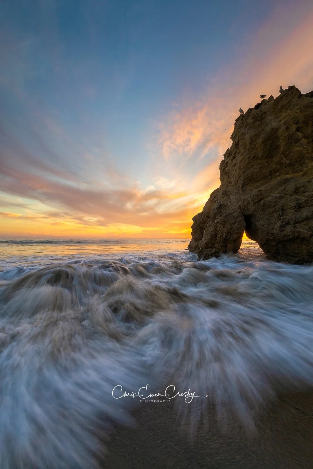 Vertical 2:3 photo of three birds silhouetted on a Malibu sea stack at sunset with misty white ocean water.