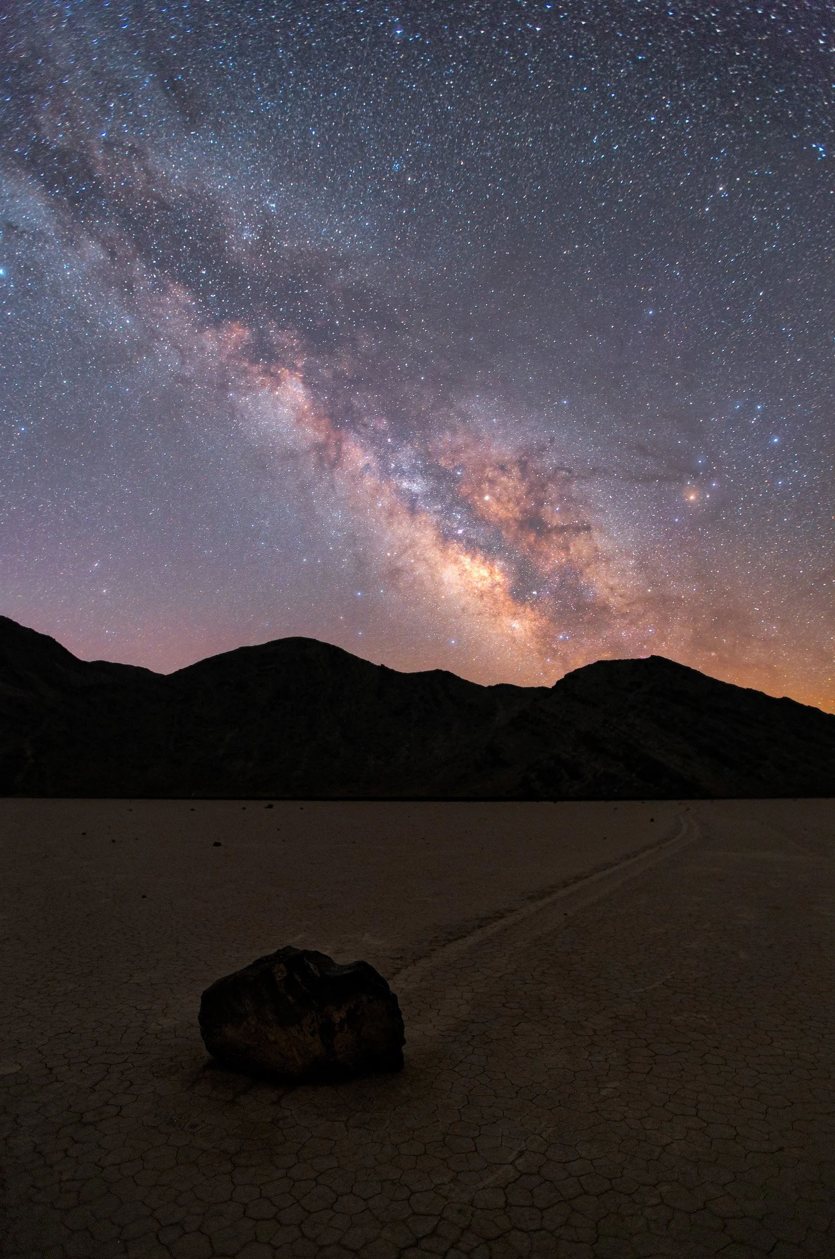 Vertical night astrophotography photograph of a sliding rock with its visible track across cracked mud beneath the Milky Way galaxy at The Racetrack Playa in Death Valley National Park by Chris Ewen Crosby.