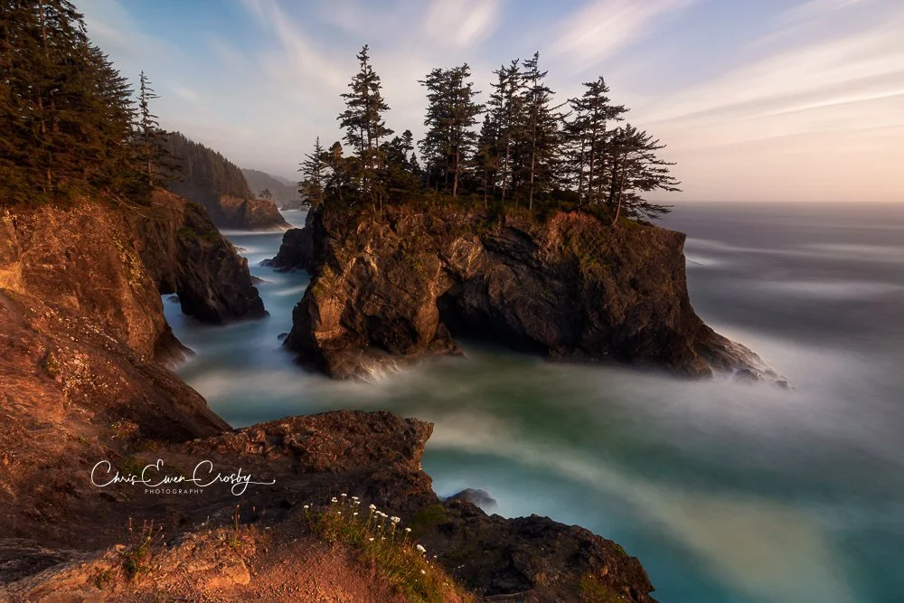 Long exposure of sea stacks and natural rock bridges on the Oregon coast with misty white water and jagged dark cliffs.