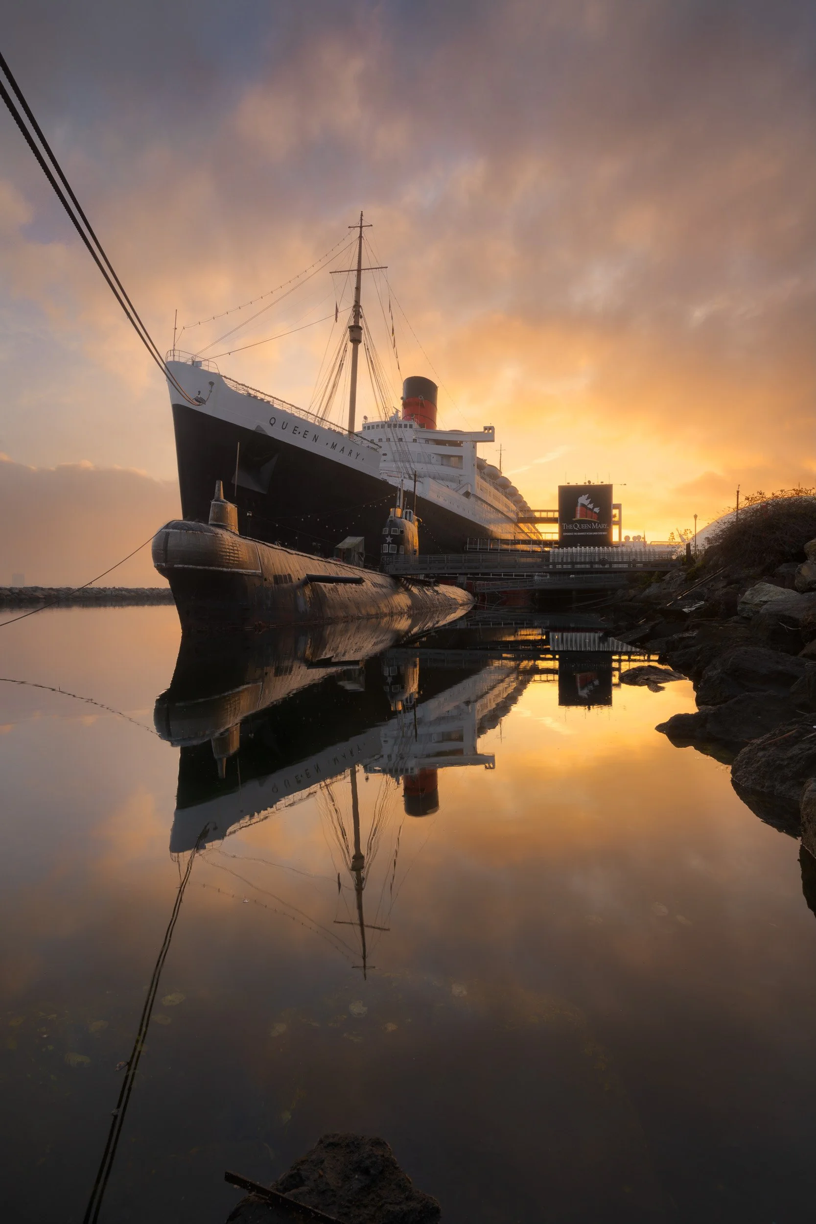 Vertical shot of the historic Queen Mary ocean liner and Soviet Scorpion submarine reflecting in still water during a golden sunrise at Long Beach Harbor by Chris Ewen Crosby.
