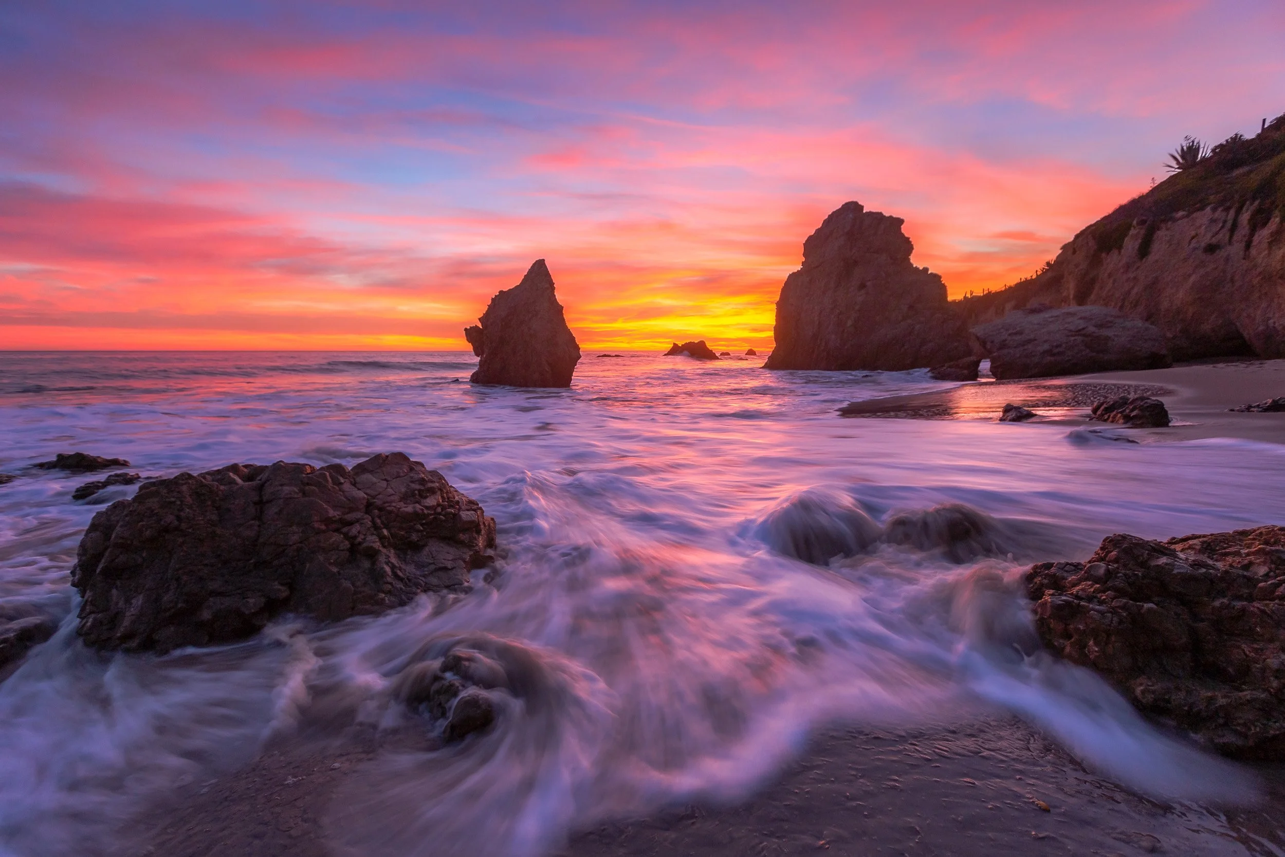 Horizontal long exposure photograph of a fiery pink and orange sunset over sea stack monoliths and rushing tide at El Matador State Beach in Malibu by Chris Ewen Crosby.