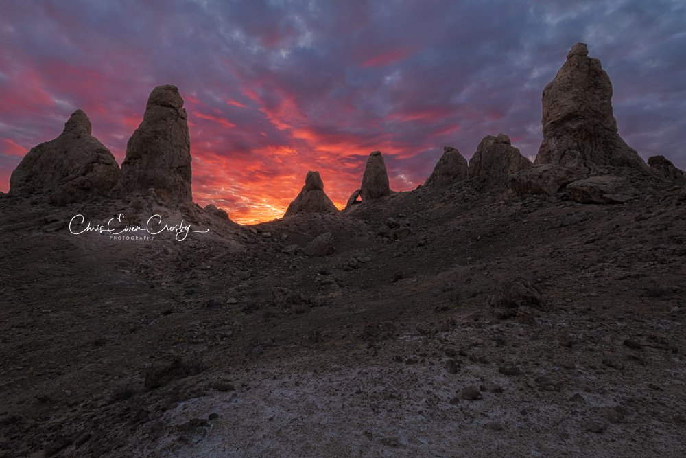 A horizontal landscape photograph of the Trona Pinnacles at sunrise, featuring jagged tufa rock formations glowing with orange light under a soft morning sky.
