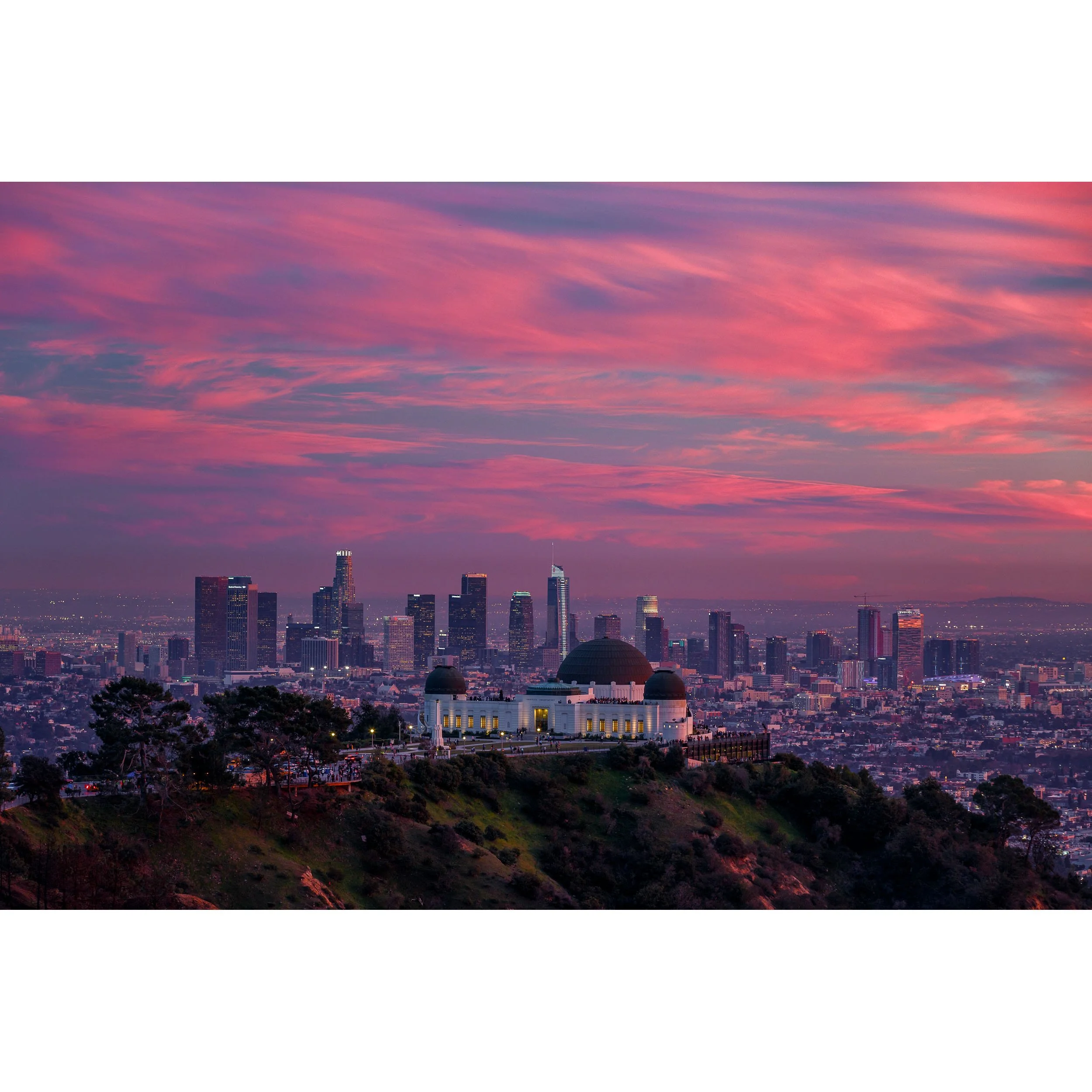 Panoramic sunset photography of the Griffith Observatory with the Downtown Los Angeles skyline beneath vibrant pink clouds.
