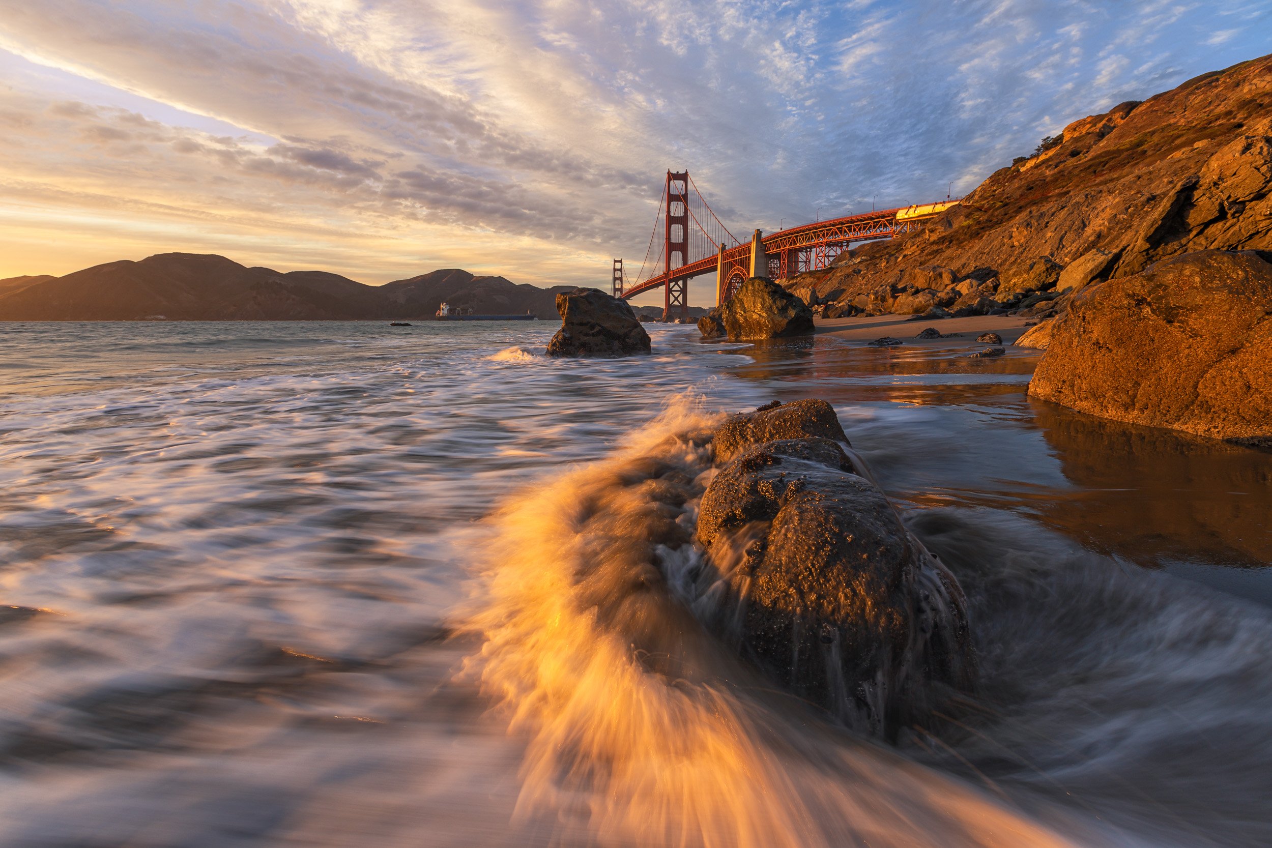 Horizontal long exposure shot of golden sunset light washing over ocean surf and rocks at Marshall's Beach with the Golden Gate Bridge in the background by Chris Ewen Crosby.