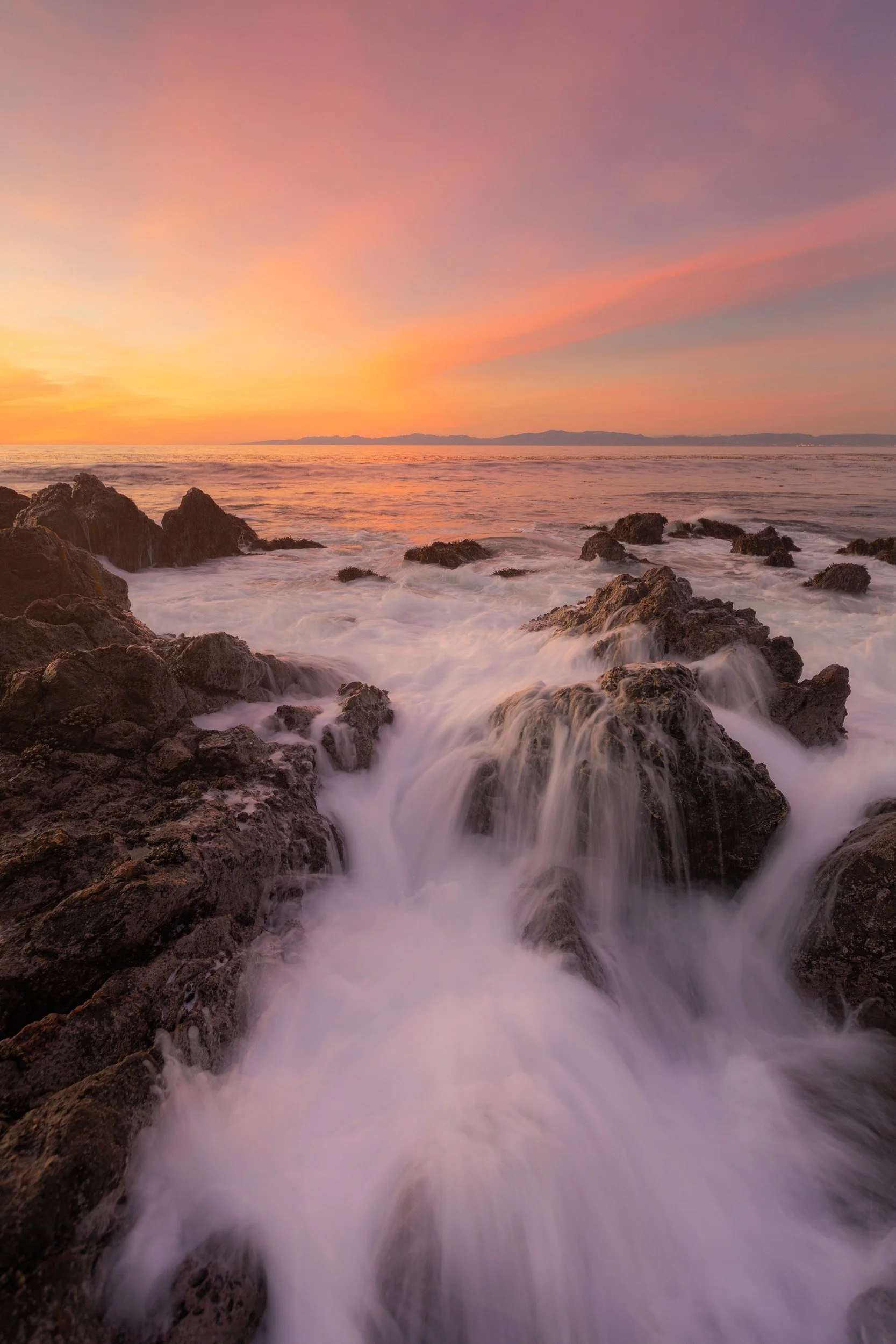 Vertical long exposure of churning ocean water cascading over coastal rocks under a soft pastel peach sunset in Palos Verdes with Catalina Island on the horizon by Chris Ewen Crosby.