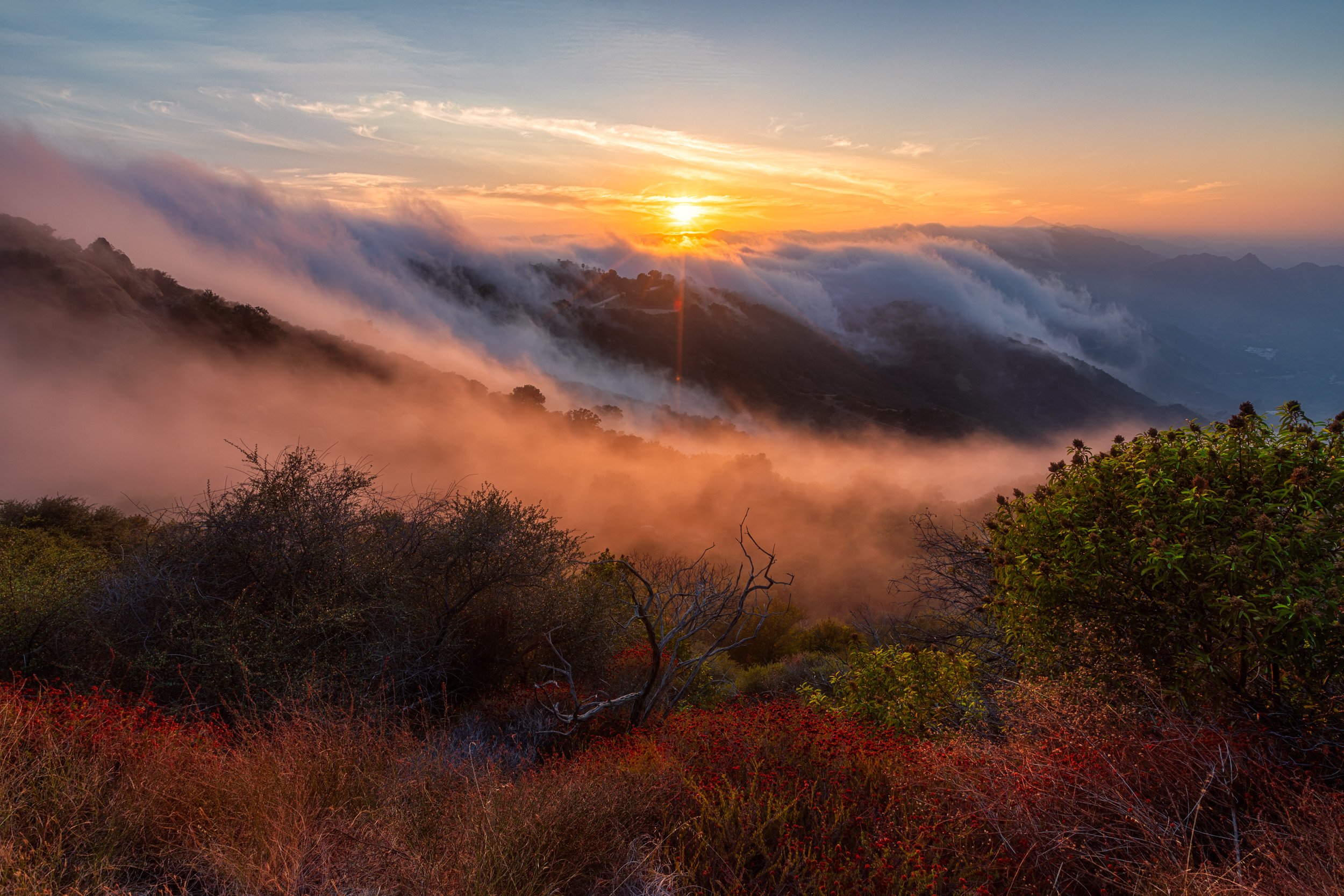 Horizontal landscape photograph of a thick ocean marine layer spilling over mountain ridges like a waterfall during a warm sunset in the Southern California mountains by Chris Ewen Crosby.