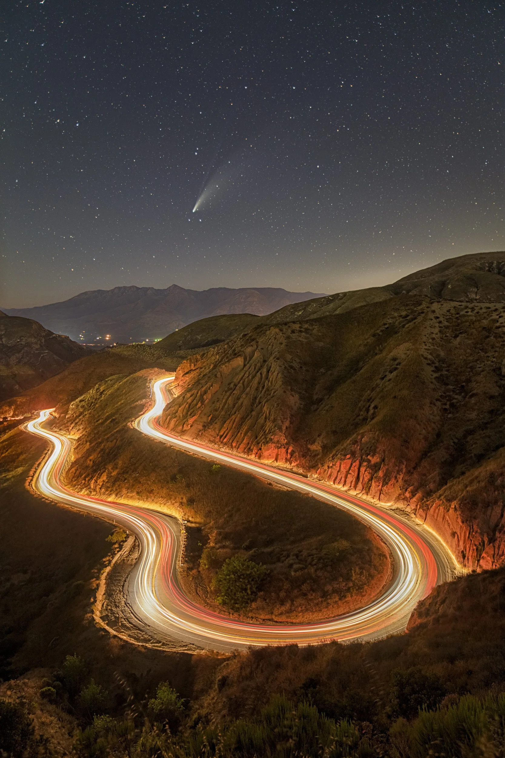 Vertical night astrophotography photograph of Comet NEOWISE glowing in the starry night sky directly above the winding S-curve headlight light trails of a mountain road by Chris Ewen Crosby.