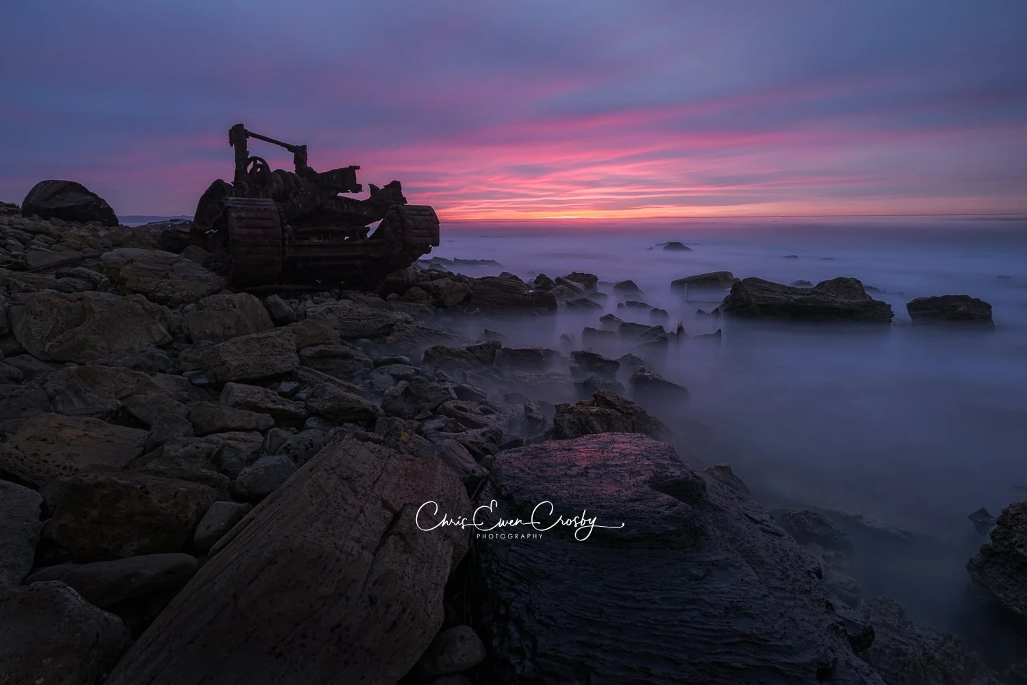 A horizontal landscape photograph titled "Forgotten Wreck" showing the rusty metal remains of a shipwreck on a rocky beach at sunset with misty, long-exposure ocean water.