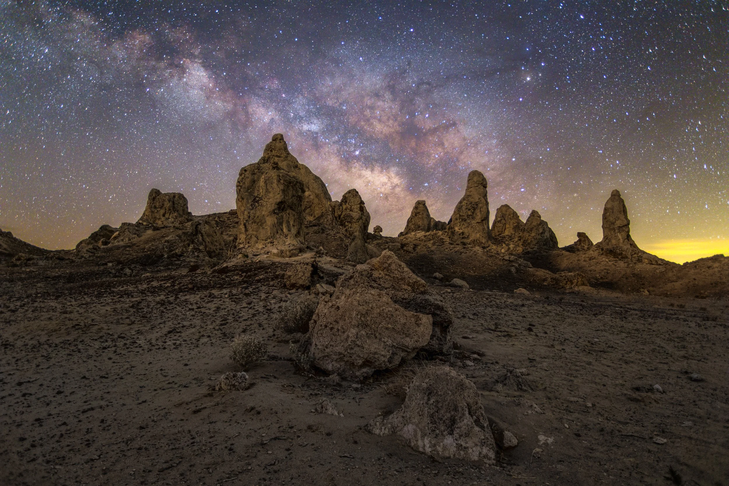 Horizontal night astrophotography photograph of a wide panoramic view of tufa rock spires spanning across the Mojave Desert beneath the arching Milky Way galaxy at Trona Pinnacles by Chris Ewen Crosby.