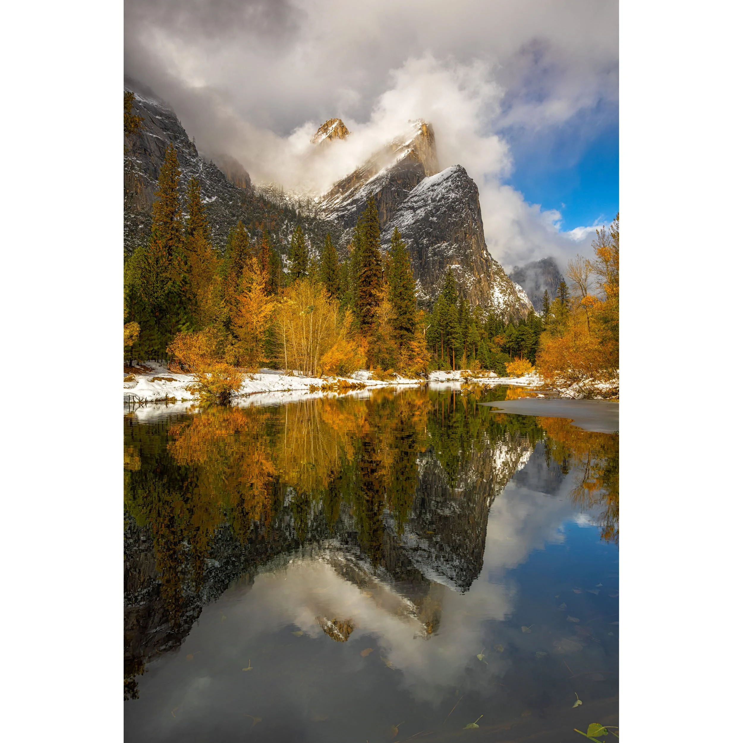 A vertical photograph in Yosemite Valley showing snowy mountains and golden fall trees reflected perfectly in a still river.