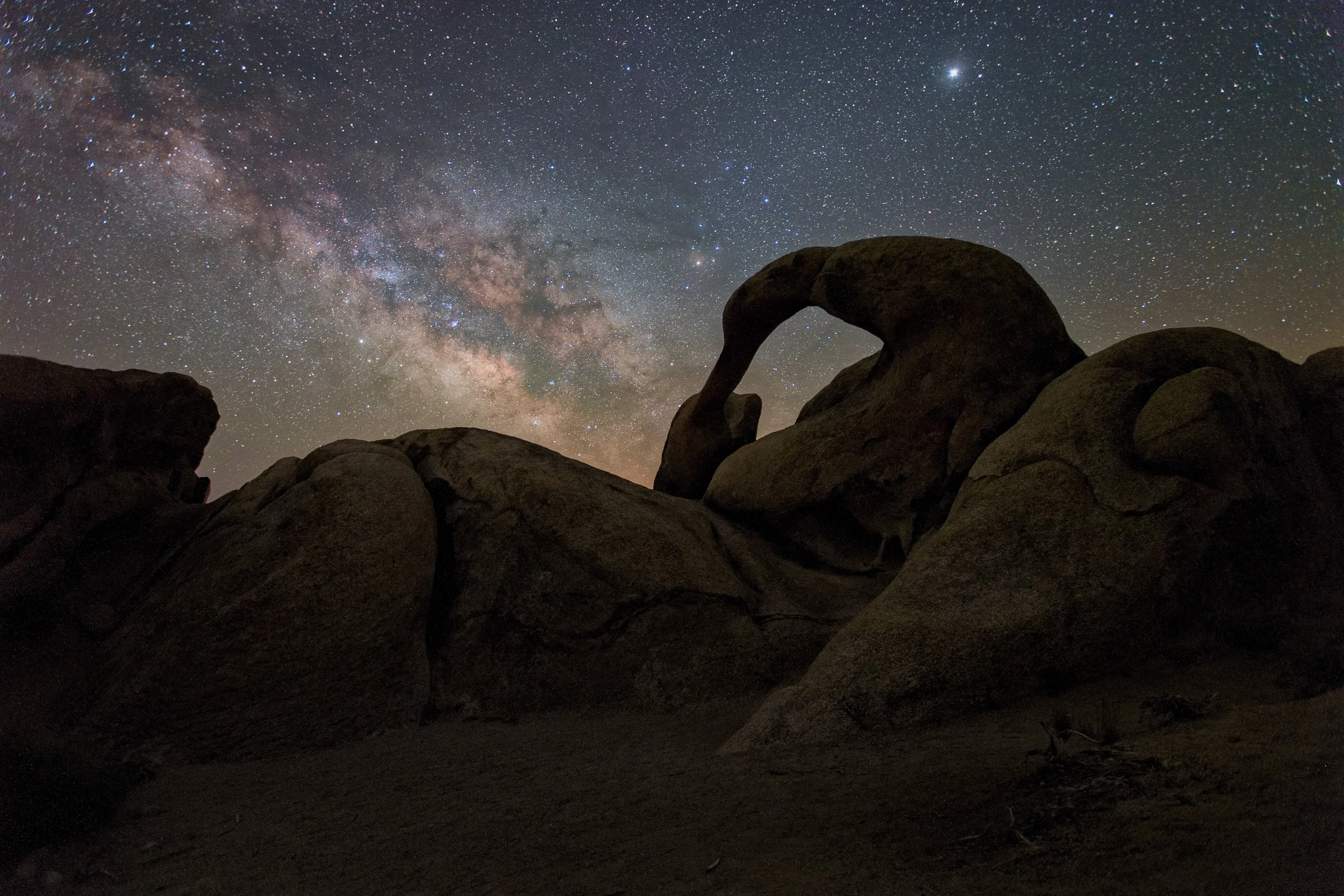 Horizontal night astrophotography photograph of the silhouetted Mobius Arch rock formation framing the glowing Milky Way galaxy core in the Alabama Hills near Lone Pine, California by Chris Ewen Crosby.