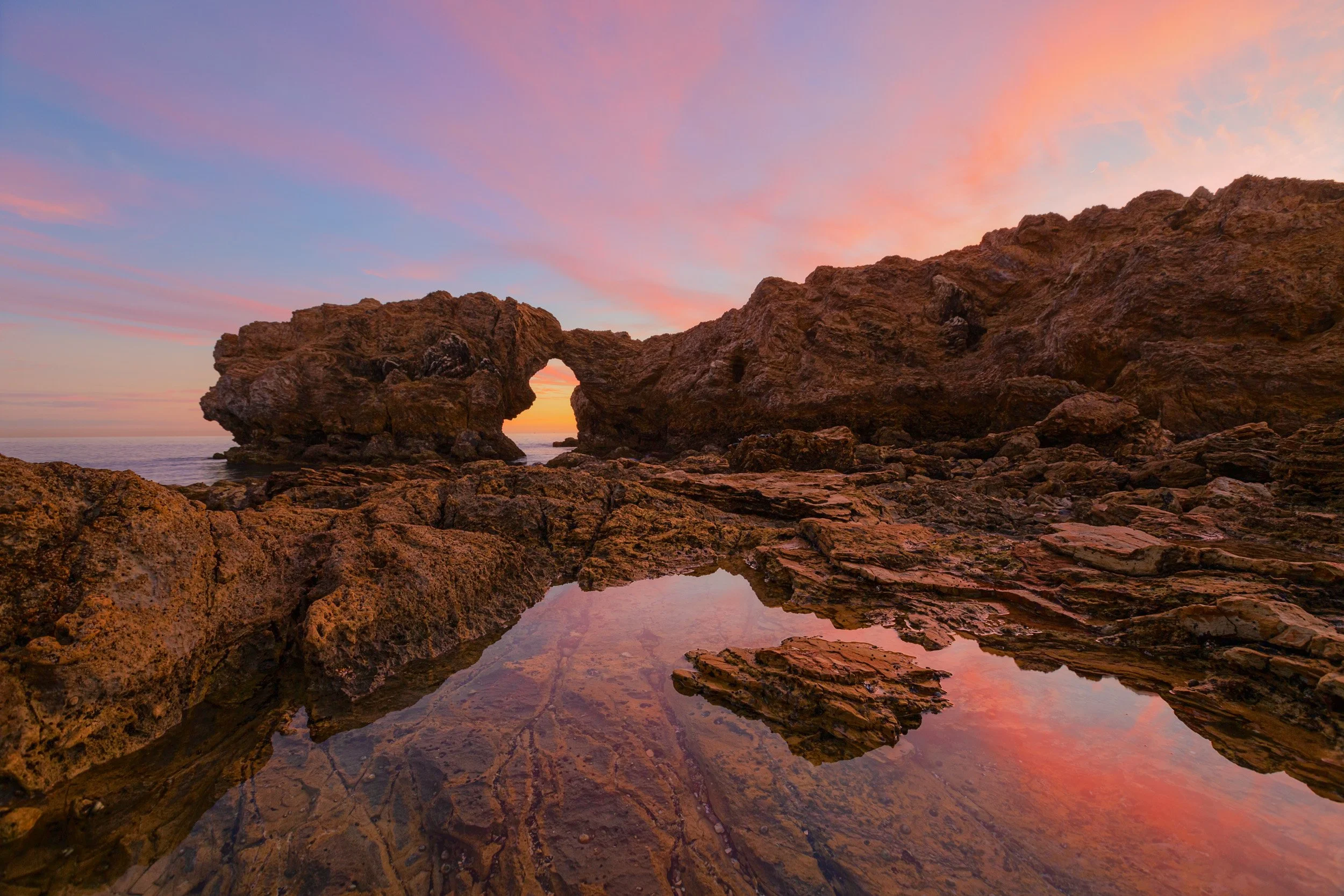 Horizontal shot of sunset light framing Arch Rock in Corona del Mar with vibrant cloud reflections in a calm tide pool by Chris Ewen Crosby.