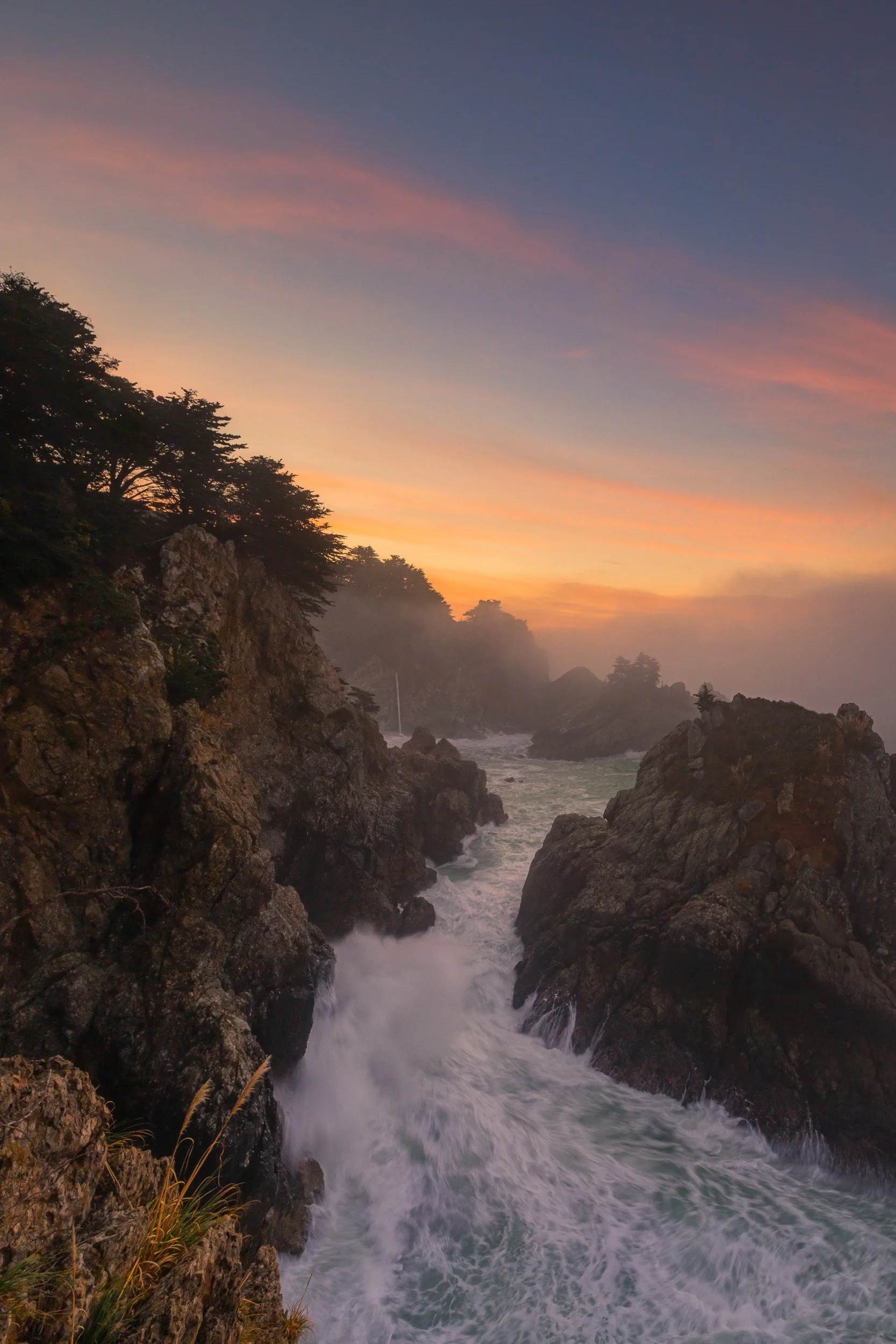 Vertical long exposure of crashing surf and misty ocean spray along the rugged Big Sur coastline with McWay Falls in the background during sunset by Chris Ewen Crosby.
