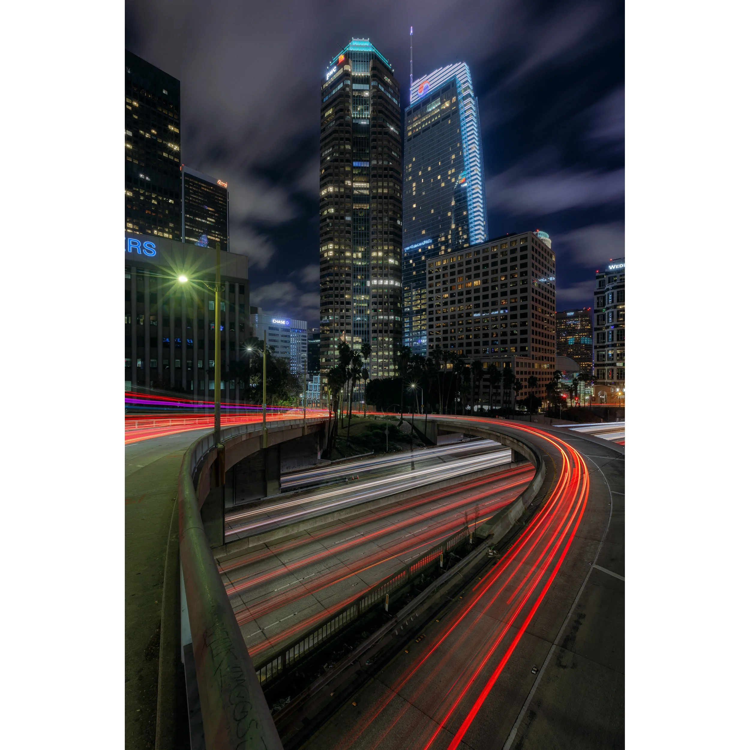 Vertical long exposure night photograph of freeway traffic light trails under illuminated Downtown Los Angeles skyscrapers.