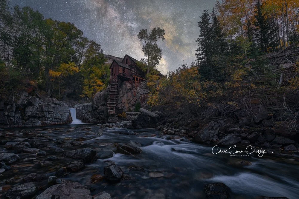 A horizontal night photograph showing the wooden Crystal Mill in Colorado perched on a cliff over a river, with the bright Milky Way galaxy stretching across the starry sky above.