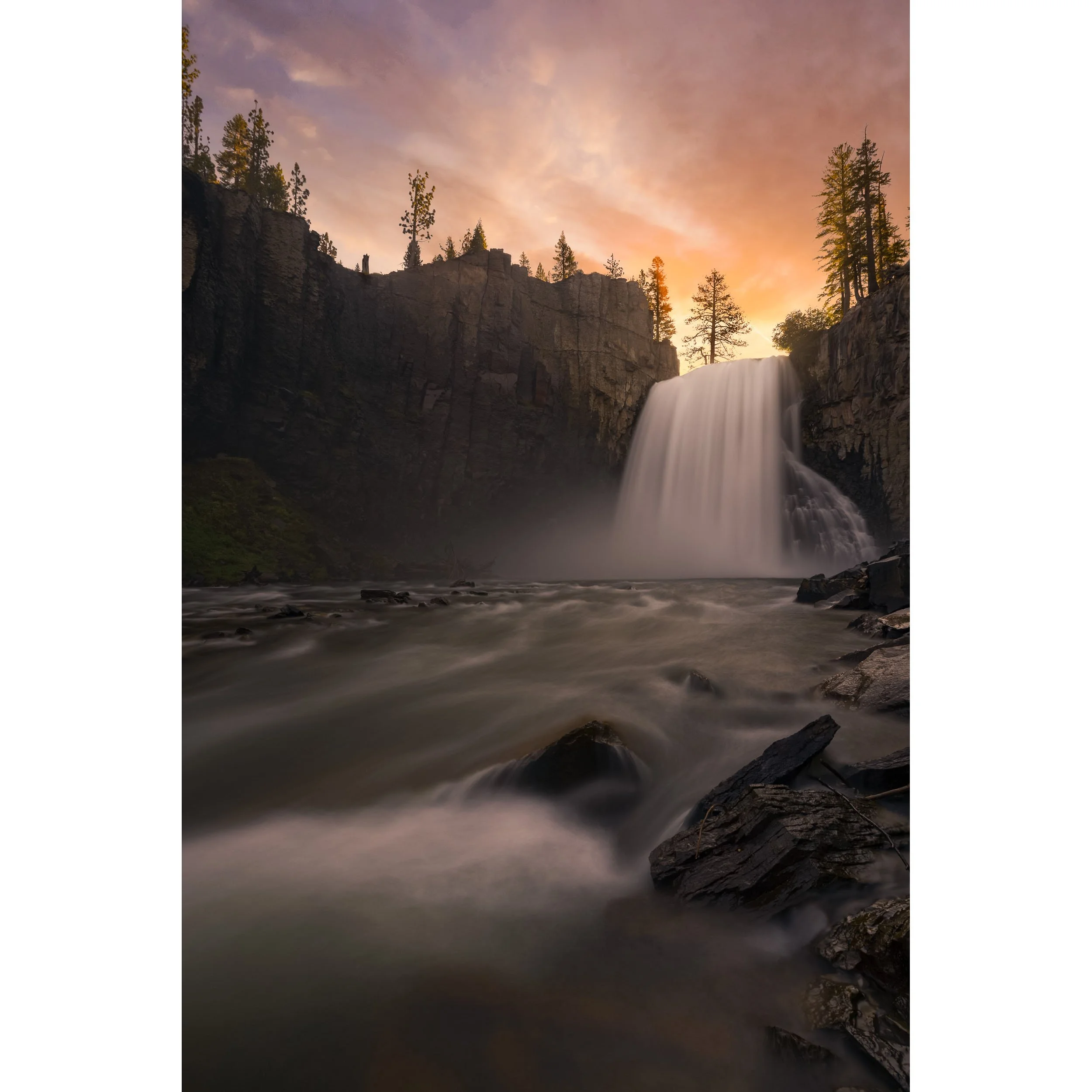 Vertical fine art landscape photograph of Rainbow Falls cascading over basalt cliffs in Mammoth Lakes under a warm sunset sky.