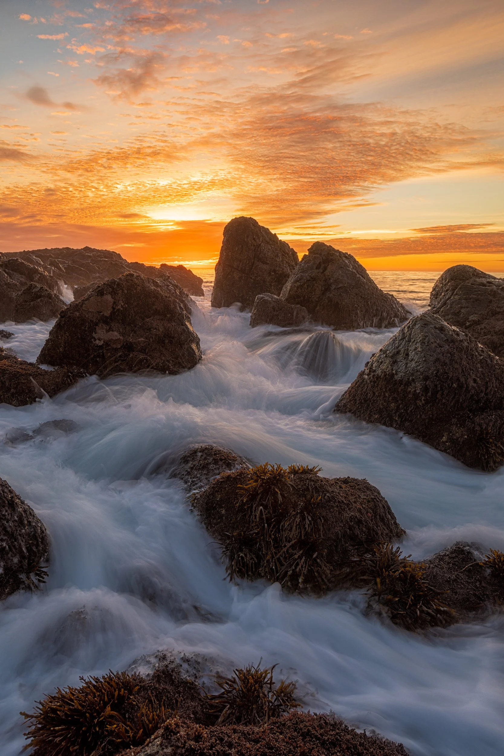 Vertical long exposure photograph of a vibrant golden sunset sky over silhouetted coastal rocks with foaming ocean water surging through the foreground in Palos Verdes by Chris Ewen Crosby.