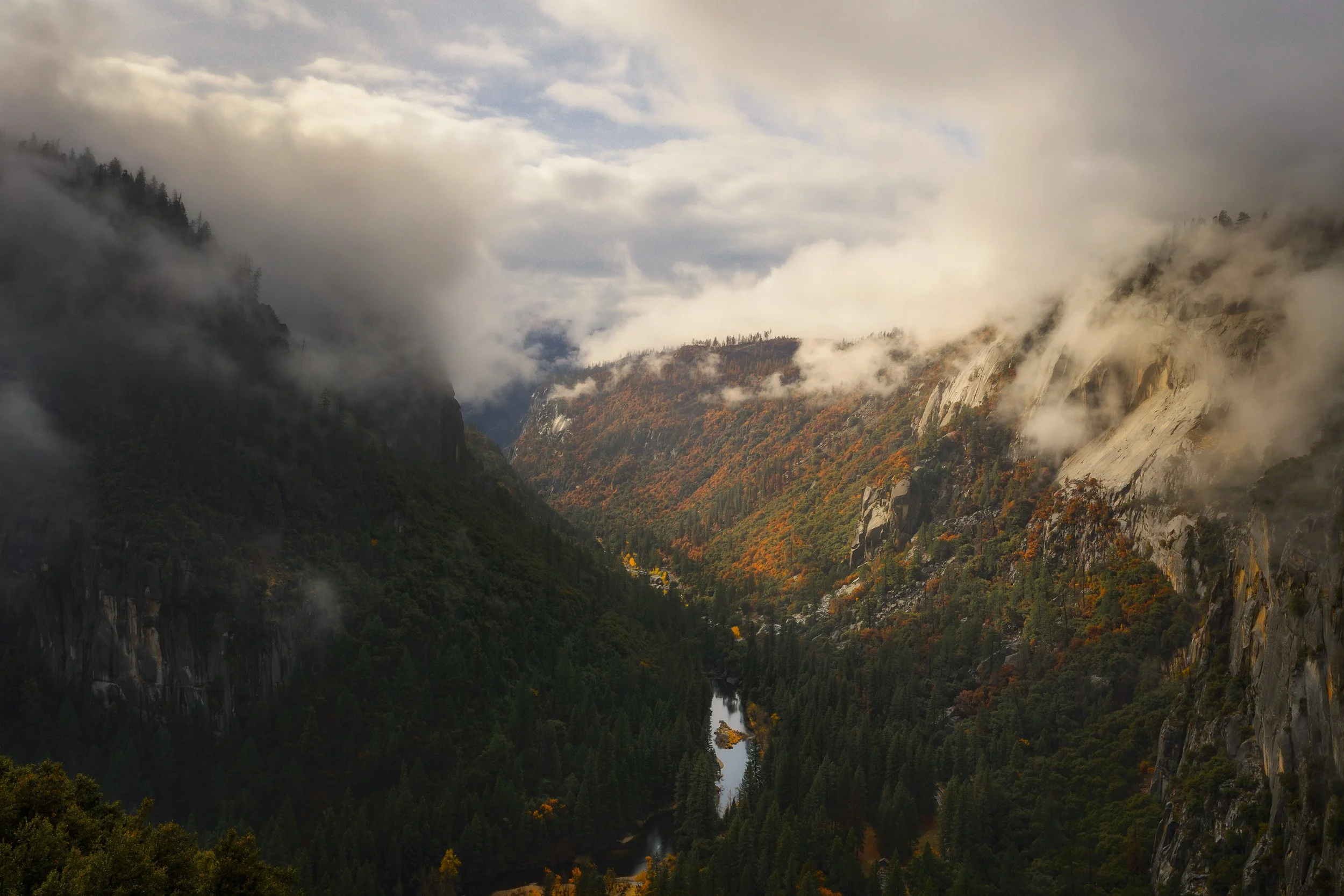 The Merced River winds through a deep Yosemite canyon flanked by low storm clouds and sunlit orange autumn foliage.