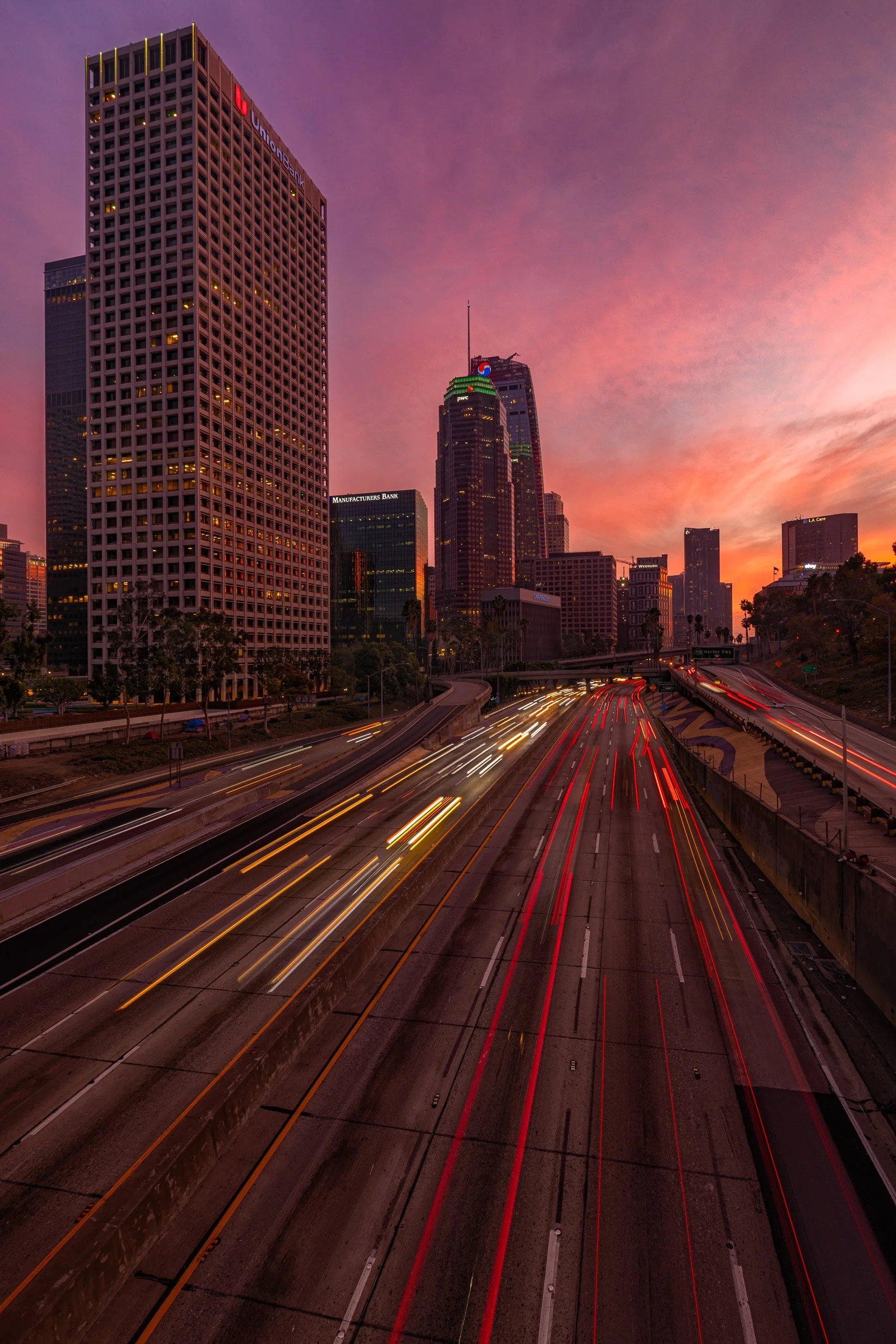 Long exposure light trails of traffic on the 110 freeway surrounded by Downtown Los Angeles skyscrapers under a purple sunset sky by Chris Ewen Crosby.