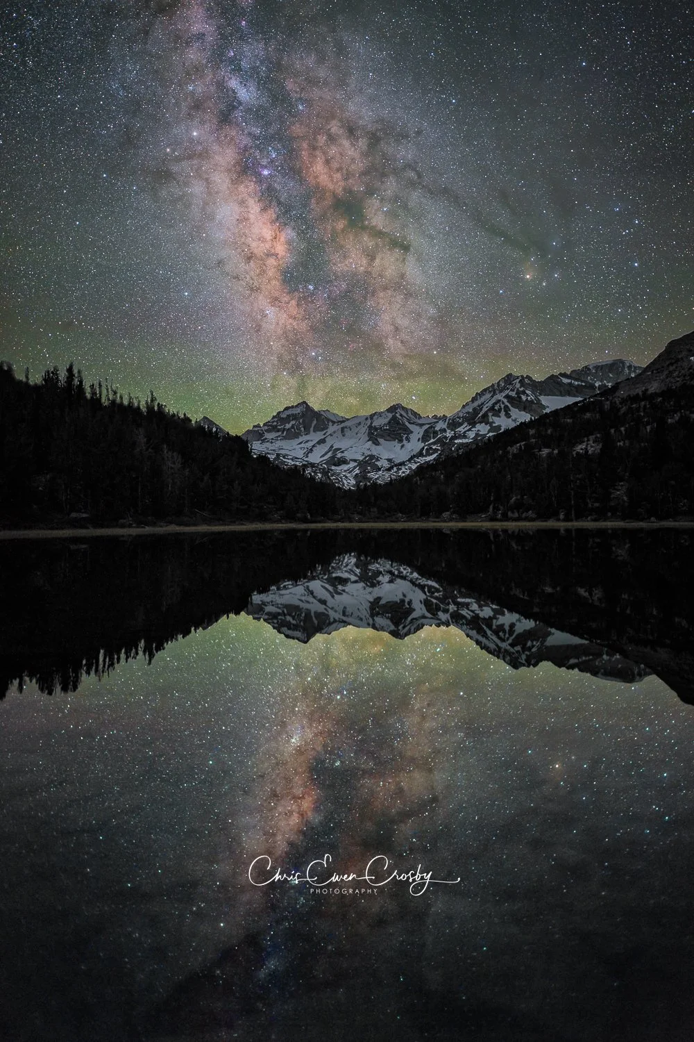 A vertical night photograph showing the bright core of the Milky Way reflected perfectly in a still alpine lake in the Sierra Nevada mountains.