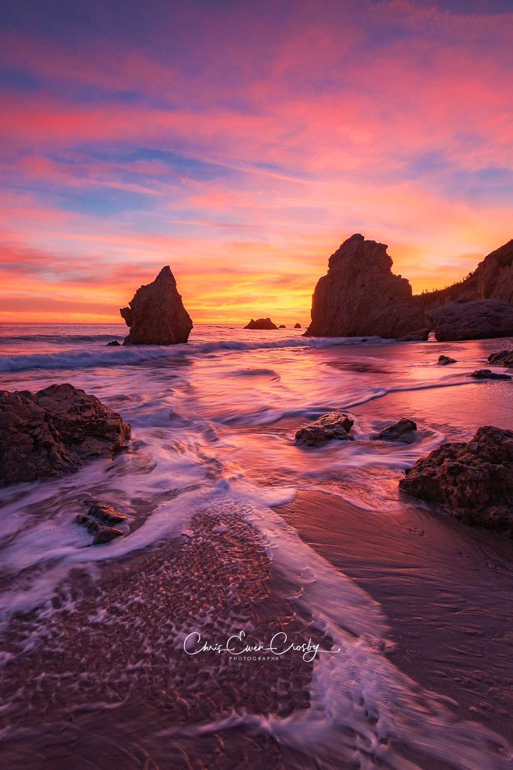 A vertical landscape photograph of El Matador State Beach at sunset, featuring dark sea stacks against a fiery orange and pink sky with water reflections.