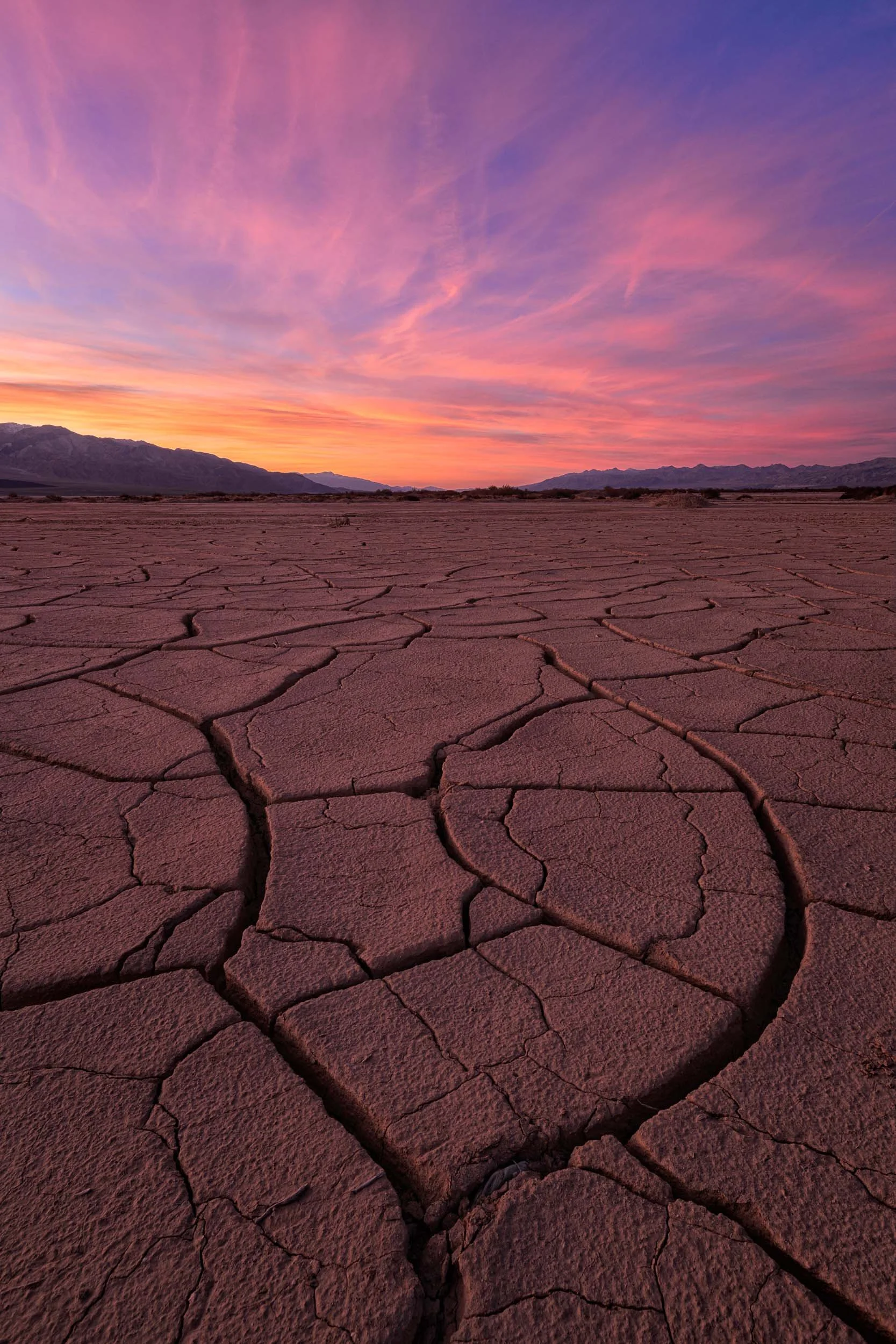 Curving cracks in a dry mud playa leading toward distant mountains under a vibrant pink and orange sunset sky in Death Valley.