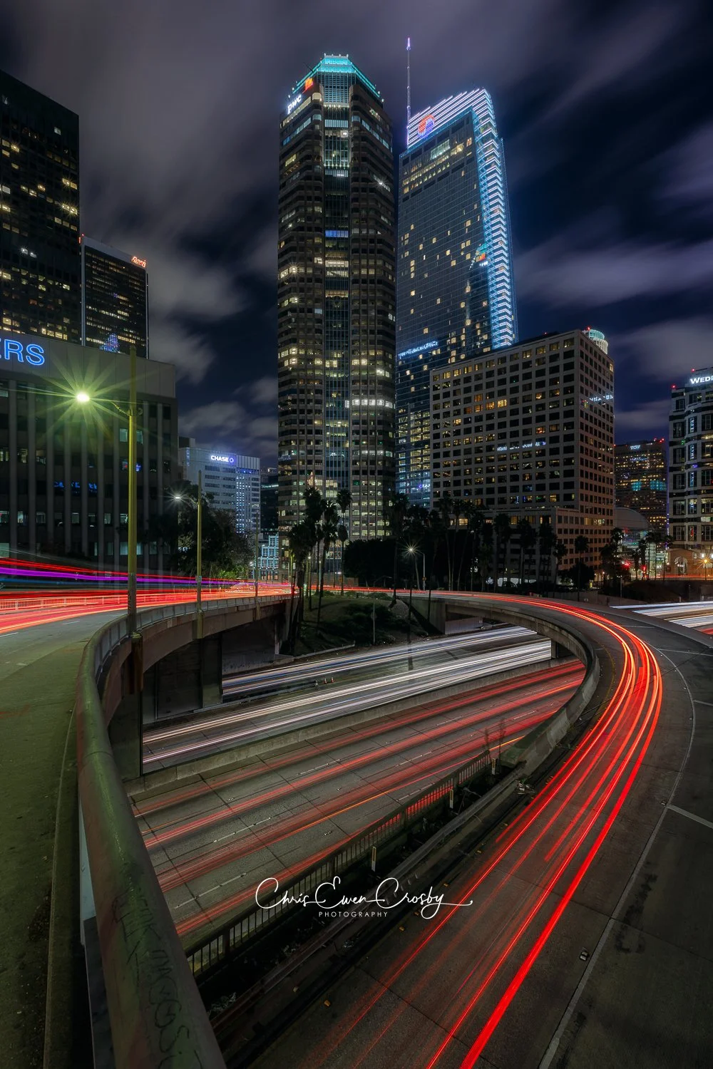 A vertical night photograph of Downtown Los Angeles featuring tall skyscrapers and glowing red and white car light trails on the freeway below.