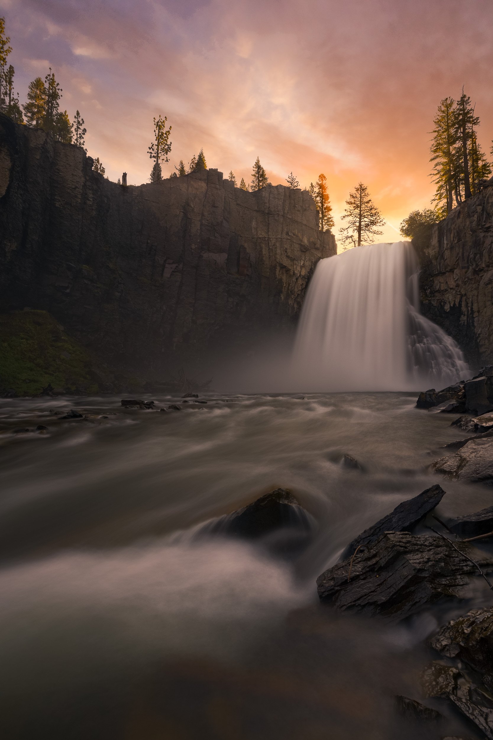 Vertical long exposure landscape photograph of Rainbow Falls plunging off basalt cliffs into the San Joaquin River under a warm sunset sky in Mammoth Lakes, California by Chris Ewen Crosby.