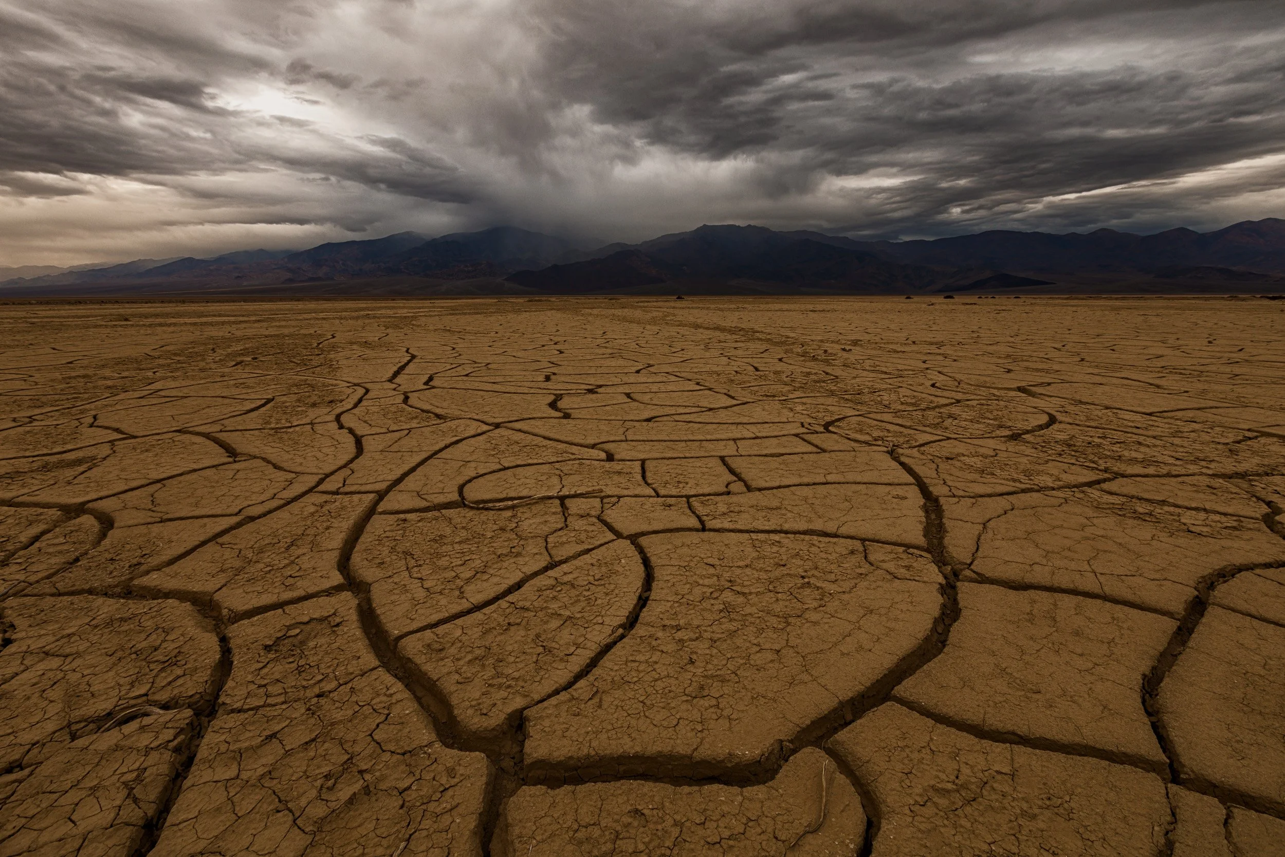 Horizontal landscape photograph of intricate cracked mud tiles on a dry desert playa leading toward distant mountains beneath a moody storm sky in Death Valley National Park by Chris Ewen Crosby.