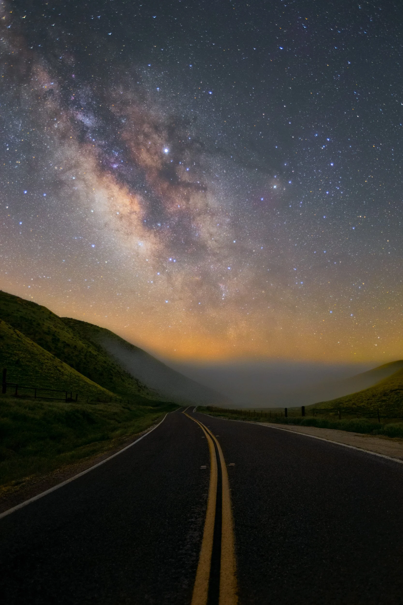 Vertical night astrophotography photograph of a two-lane asphalt road leading through rolling green hills toward low fog and the Milky Way galaxy in California Valley by Chris Ewen Crosby.