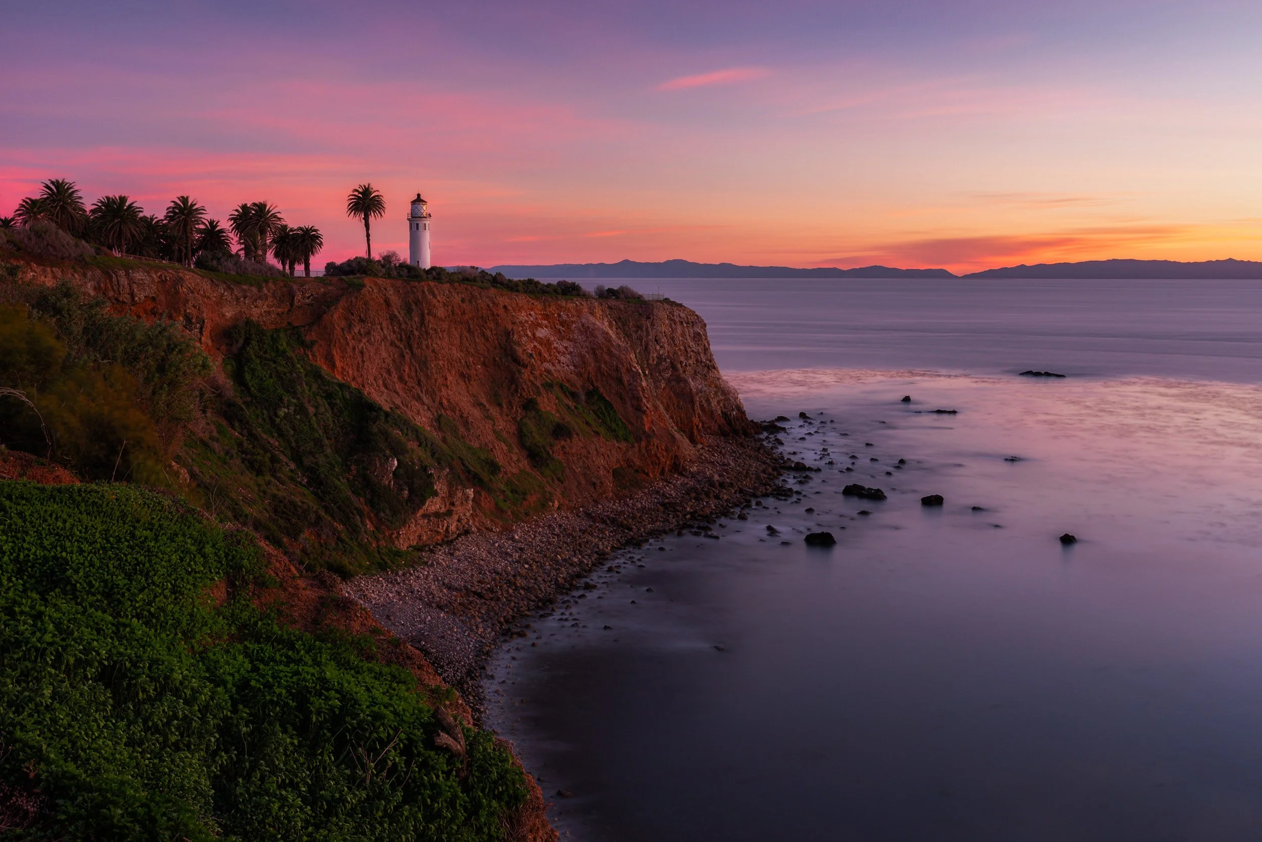 Horizontal long exposure photograph of the historic Point Vicente Lighthouse perched on coastal cliffs over smooth ocean waters during a pink sunset in Palos Verdes by Chris Ewen Crosby.