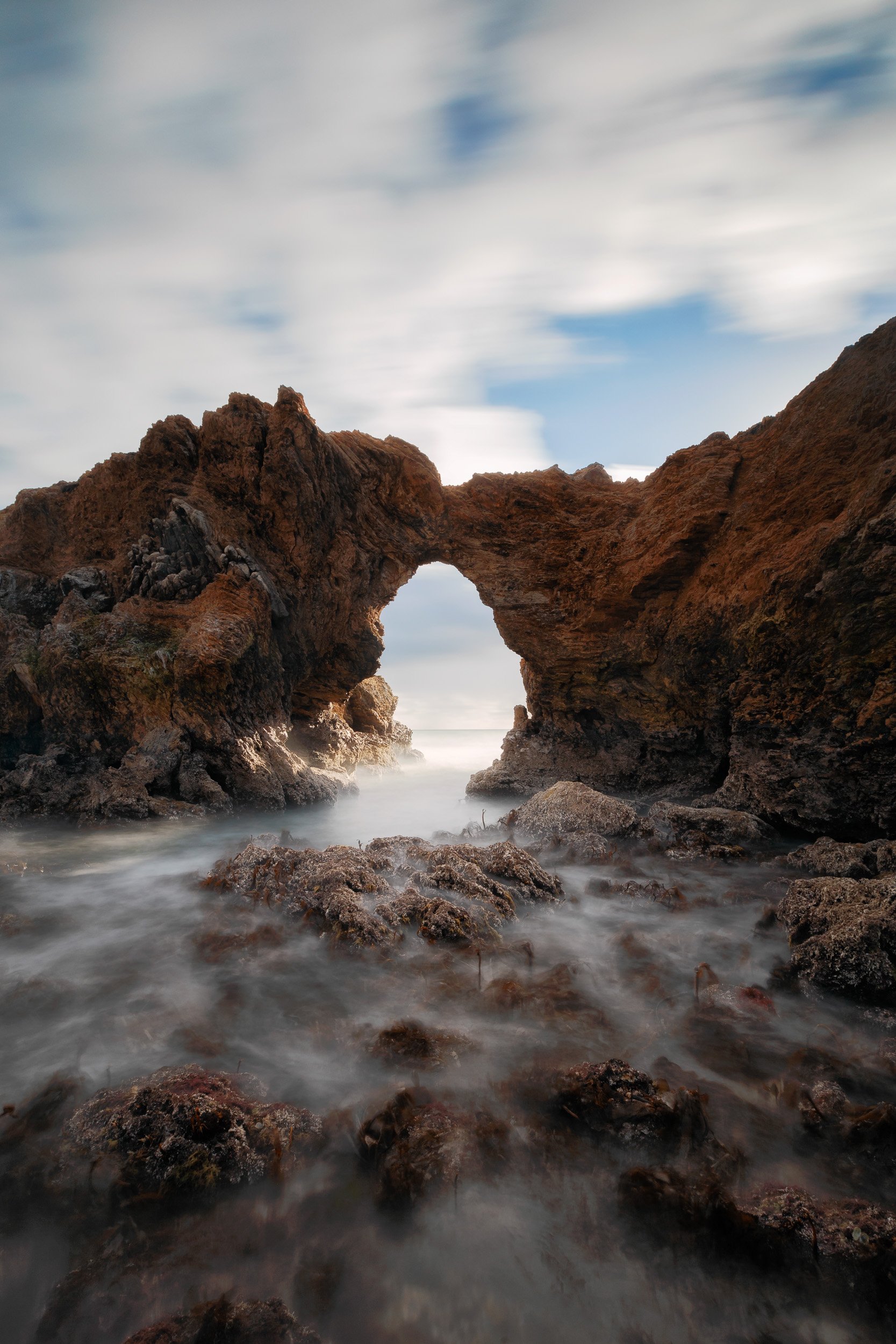 Vertical long exposure of ocean tide flowing smoothly beneath a dramatic natural sea arch rock formation at Corona del Mar State Beach by Chris Ewen Crosby.