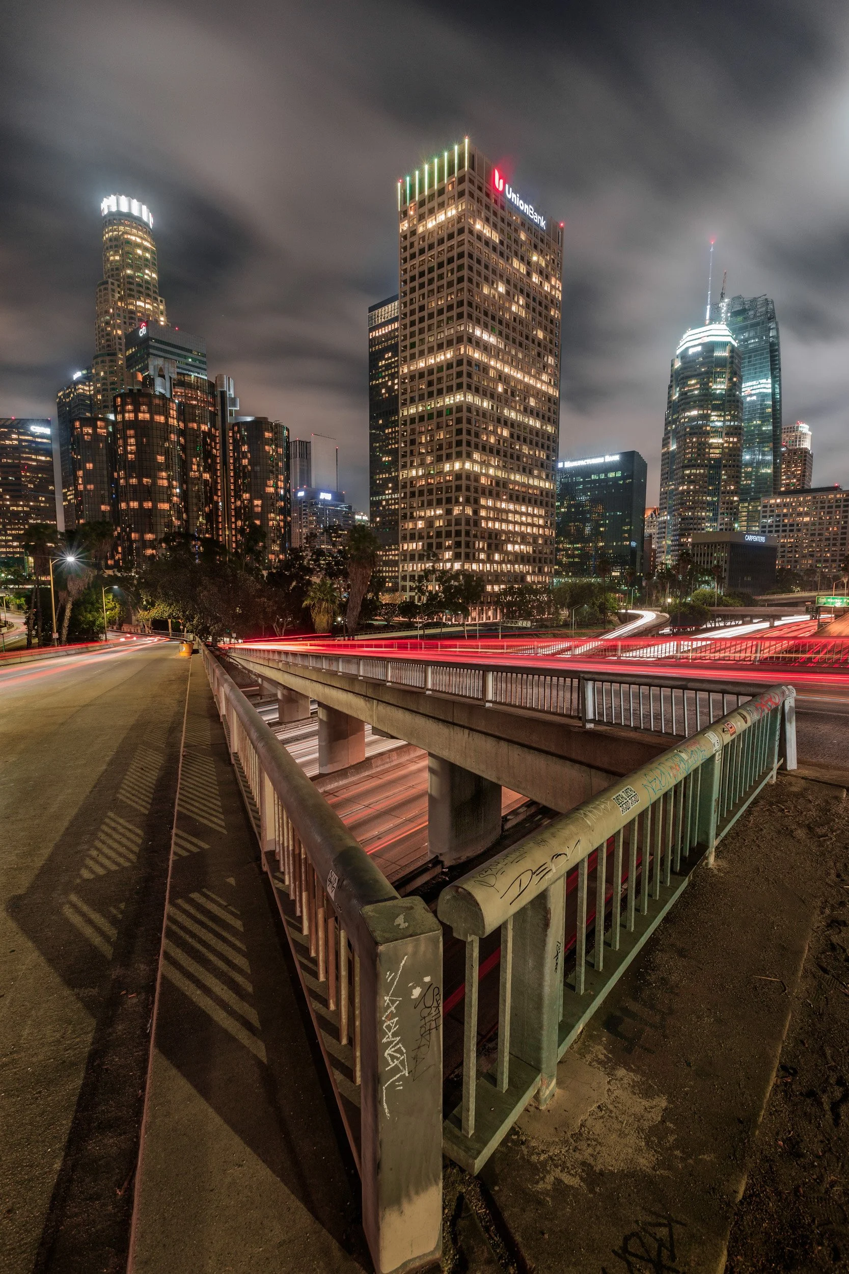 Nighttime long exposure of traffic light trails beneath an overpass looking toward Union Bank Plaza and the Downtown Los Angeles skyline by Chris Ewen Crosby.