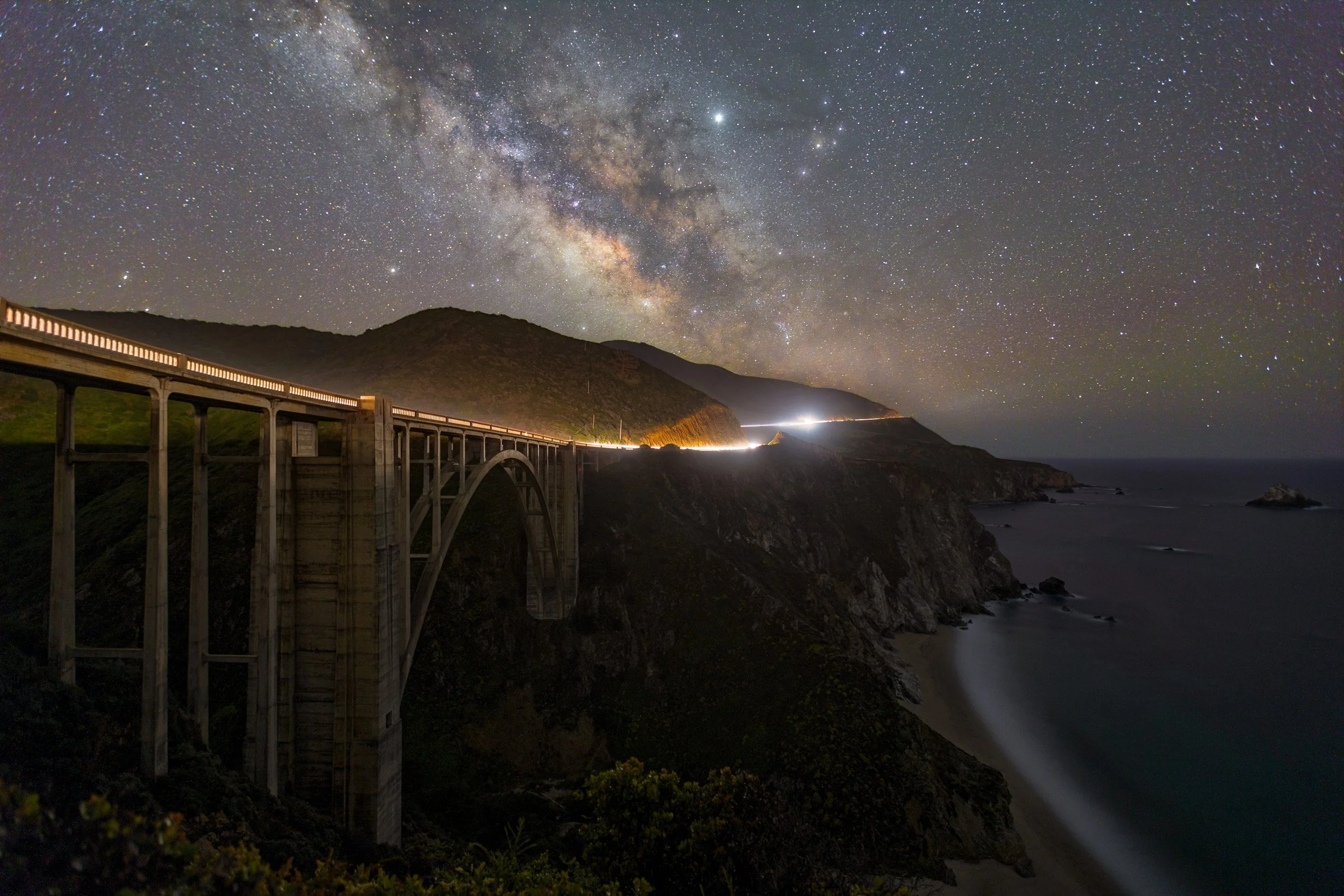 Horizontal long-exposure photograph of the Milky Way galaxy arching over Bixby Creek Bridge along the rugged Big Sur coastline at night.