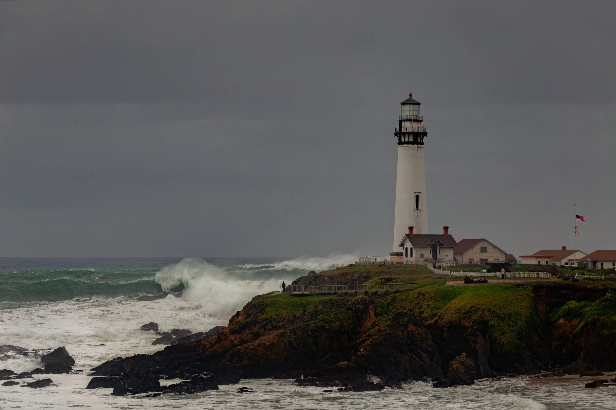 Horizontal shot of large storm waves crashing against rocky cliffs below the Pigeon Point Lighthouse under a moody overcast sky by Chris Ewen Crosby.