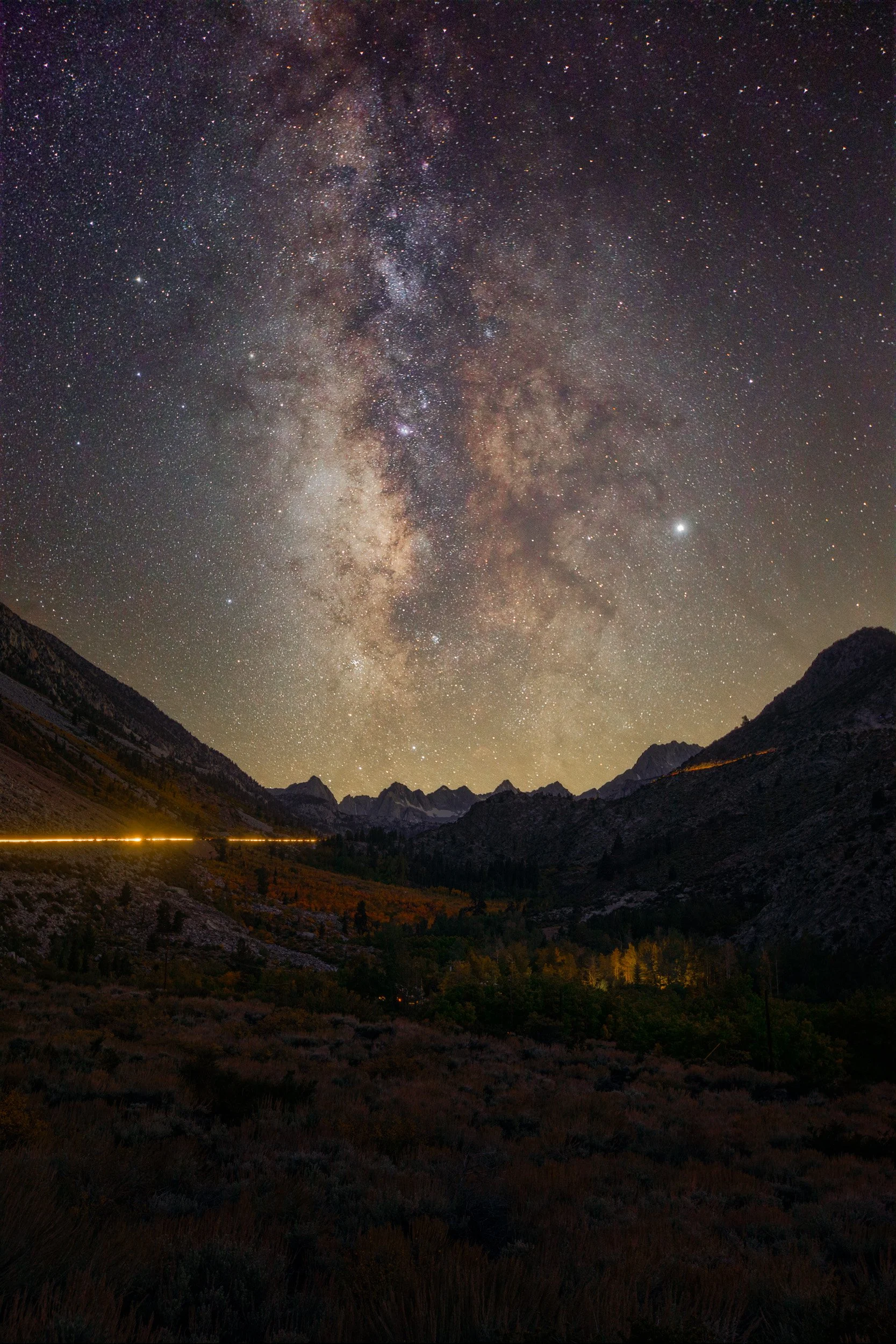 Vertical night astrophotography photograph of the Milky Way core towering above rugged mountain peaks and winding car light trails in Bishop Creek Canyon, California by Chris Ewen Crosby.