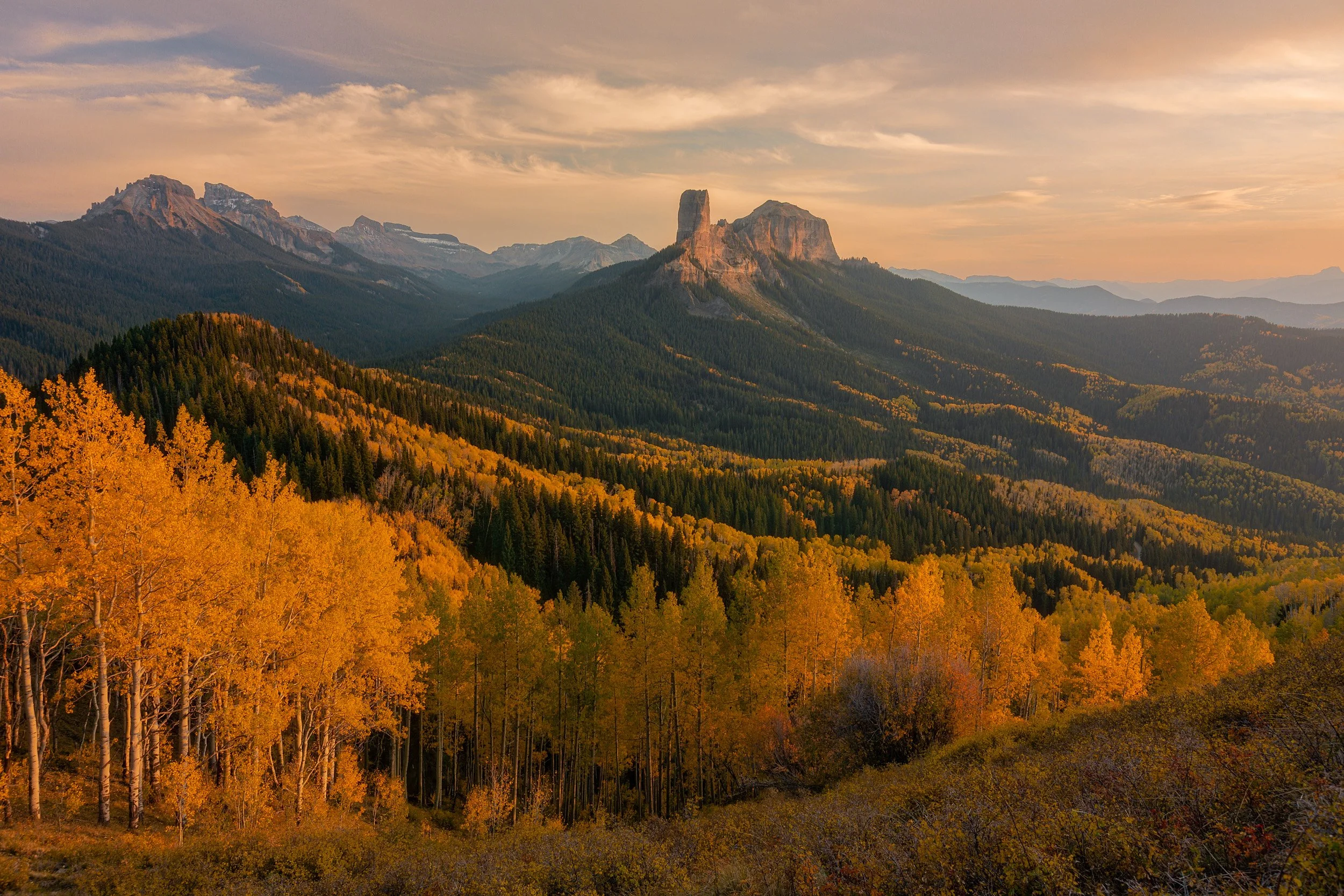 Horizontal landscape photograph of golden autumn aspen forests sweeping toward the iconic geological spire of Chimney Rock in the San Juan Mountains of Colorado by Chris Ewen Crosby.