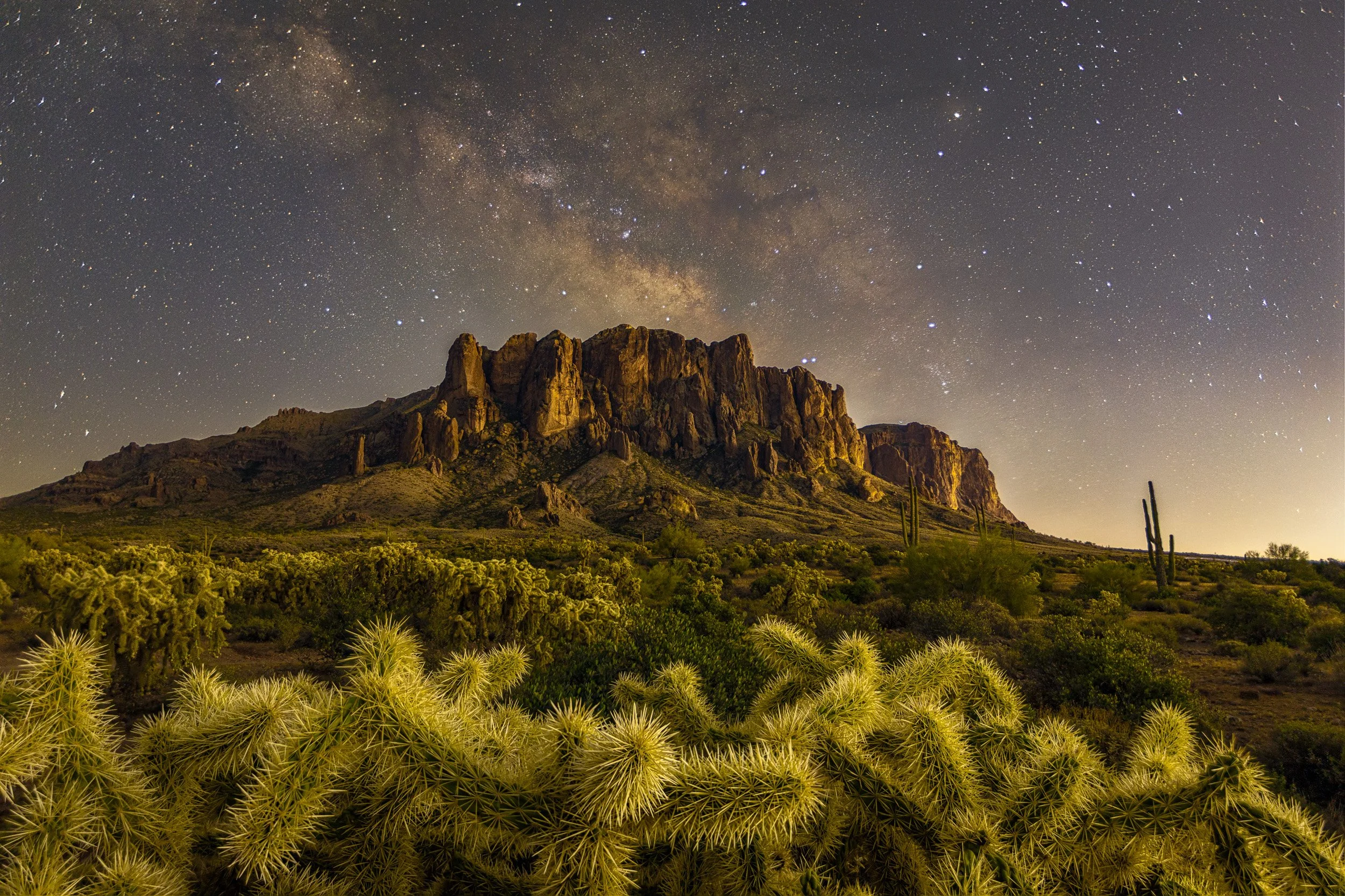 Horizontal night astrophotography photograph of the Milky Way galaxy stretching over the Superstition Mountains with glowing teddy bear cholla cactus in the foreground near Apache Junction, Arizona by Chris Ewen Crosby.