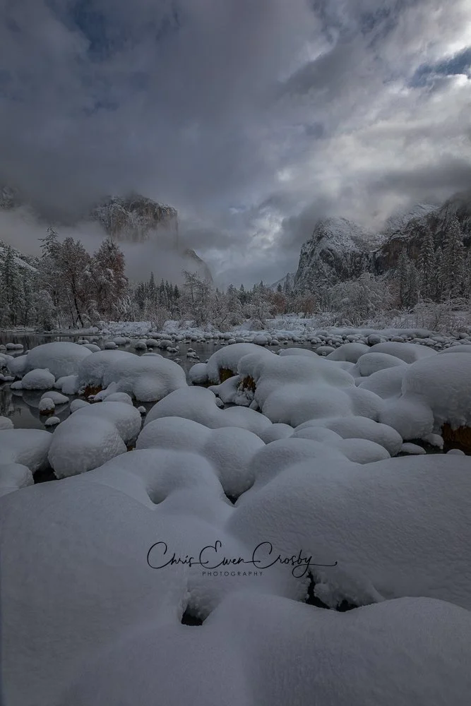 Horizontal landscape photograph of Yosemite Valley in winter. Snow-dusted rocks frame a dark river with misty granite cliffs and pine trees in the background.