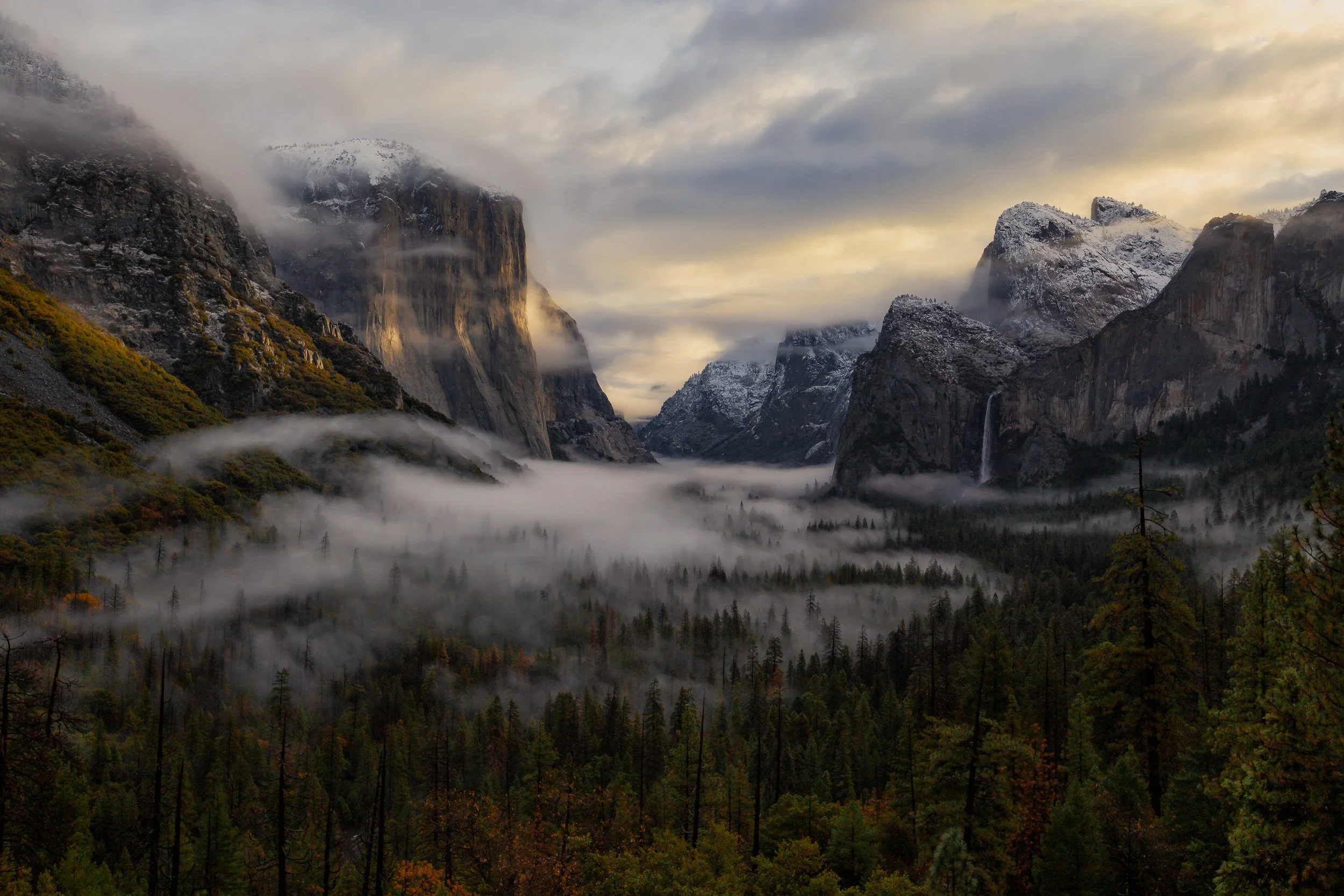 Horizontal landscape photograph of Yosemite Valley at sunset with low-lying valley fog, snow-dusted granite cliffs, and golden light hitting El Capitan.