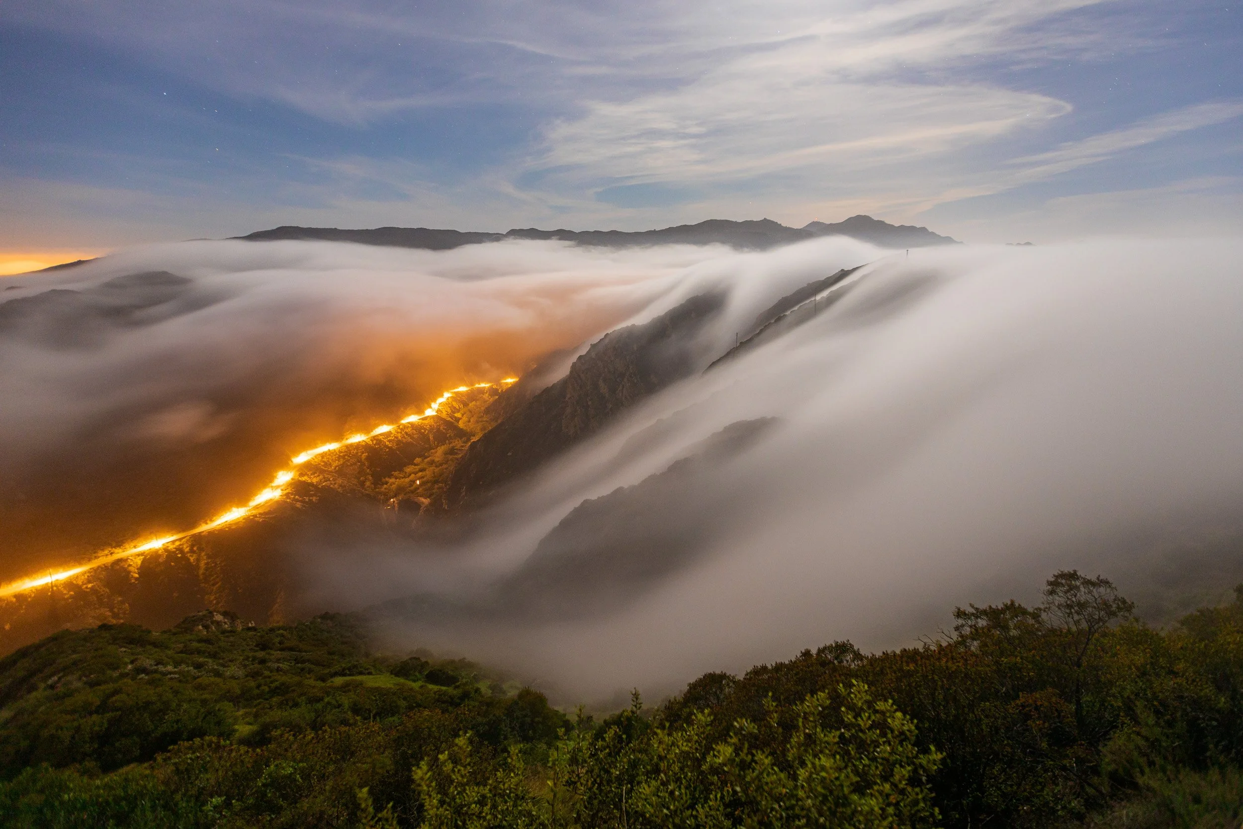 Horizontal night long exposure landscape photograph of thick coastal fog cascading over Santa Monica Mountain ridges like a waterfall while glowing car light trails illuminate a canyon below by Chris Ewen Crosby.