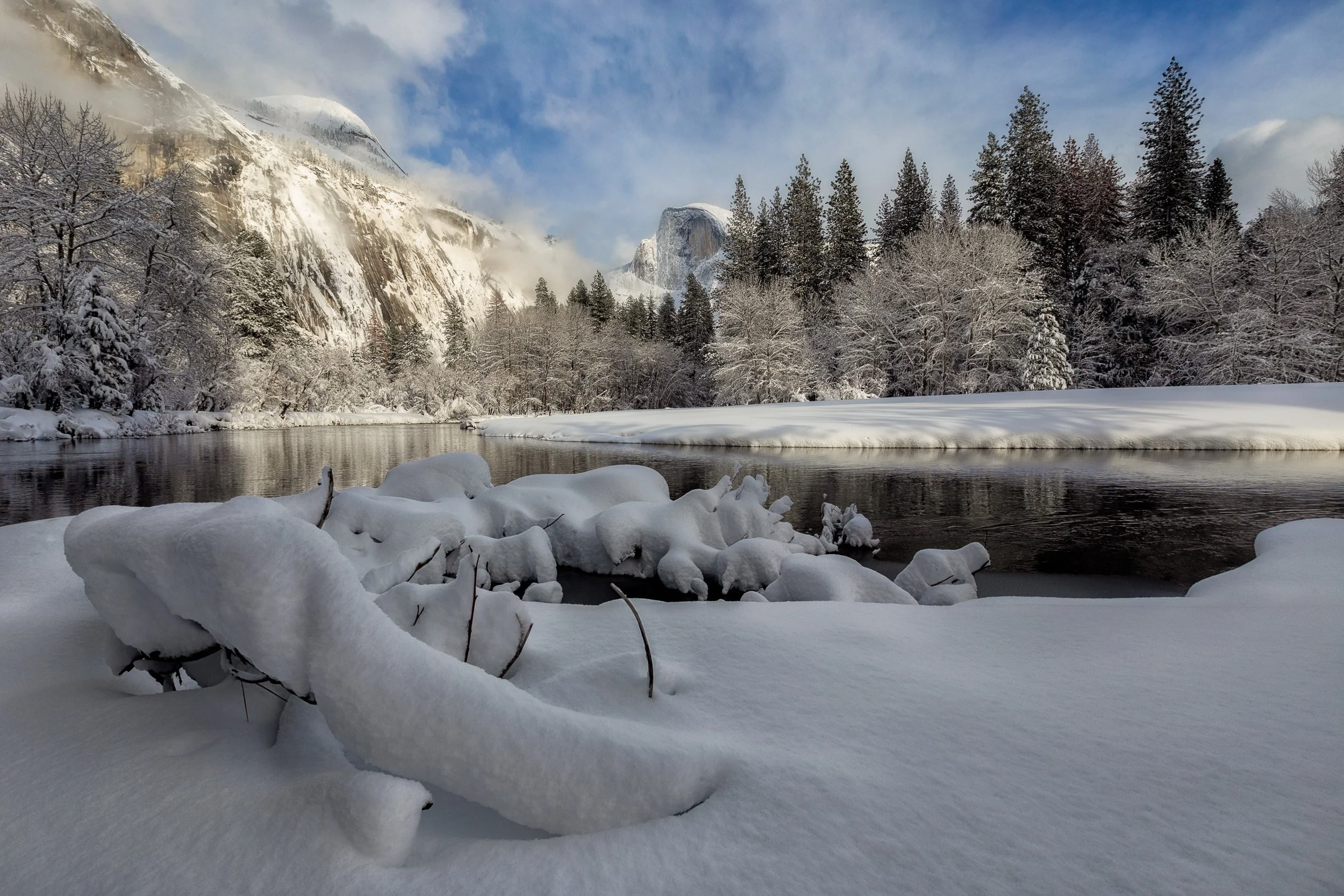 Horizontal landscape photograph of snow-laden branches along the tranquil Merced River with a snow-capped Half Dome rising in the winter fog in Yosemite National Park by Chris Ewen Crosby.
