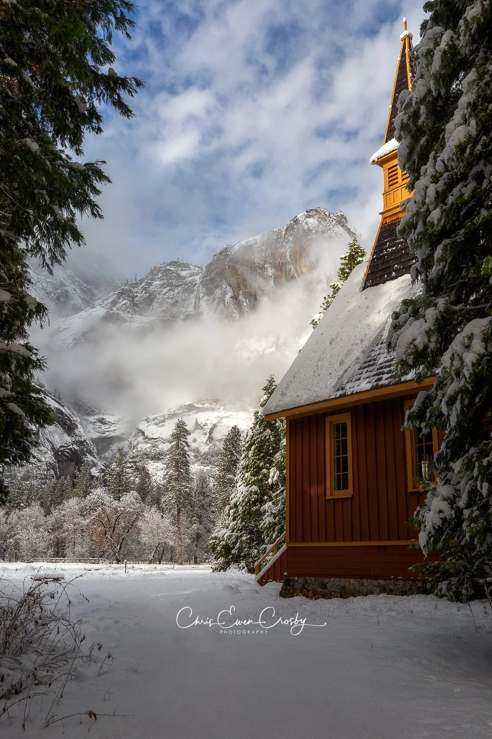 A vertical winter photograph titled "Snow Chapel" showing the historic red Yosemite Chapel covered in snow with sunlight hitting the mountains and clouds in the background.