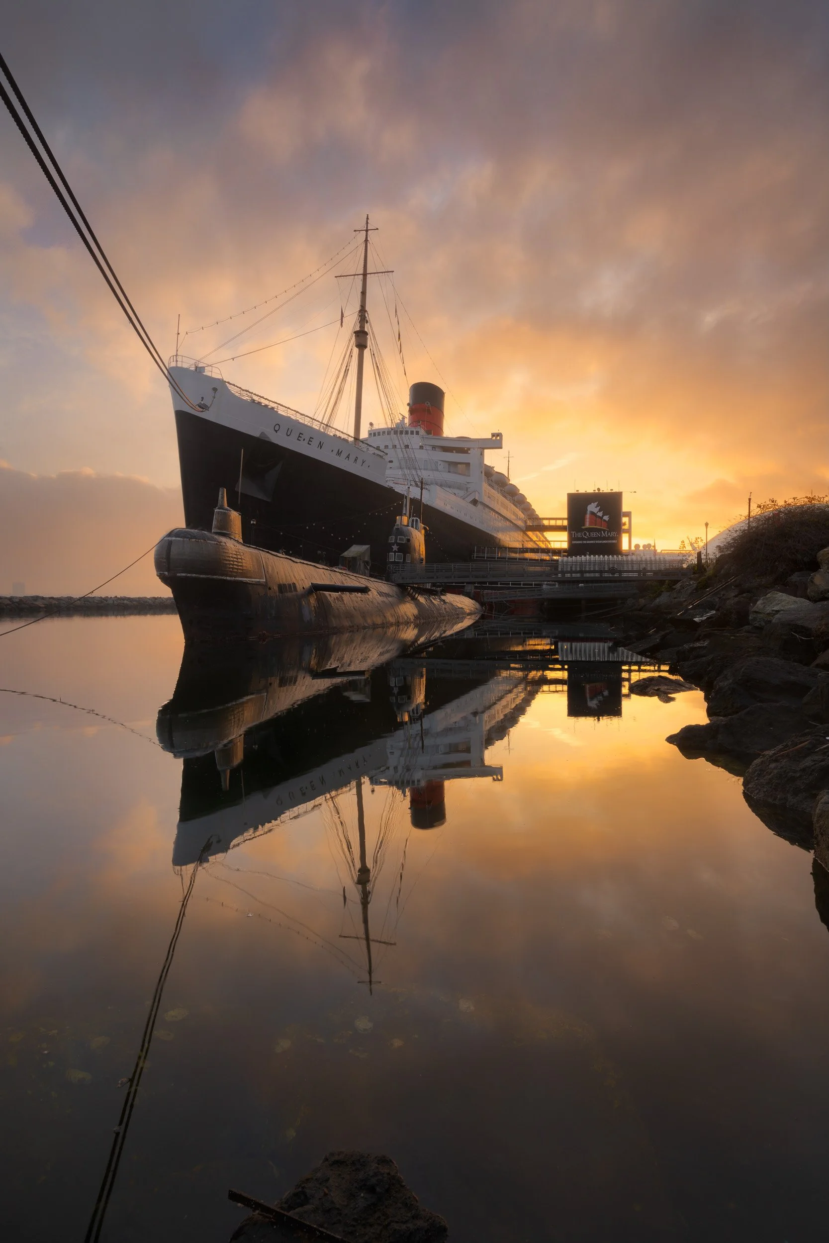 Golden sunset reflection of the RMS Queen Mary ocean liner and Scorpion submarine in calm Long Beach harbor water by Chris Ewen Crosby.