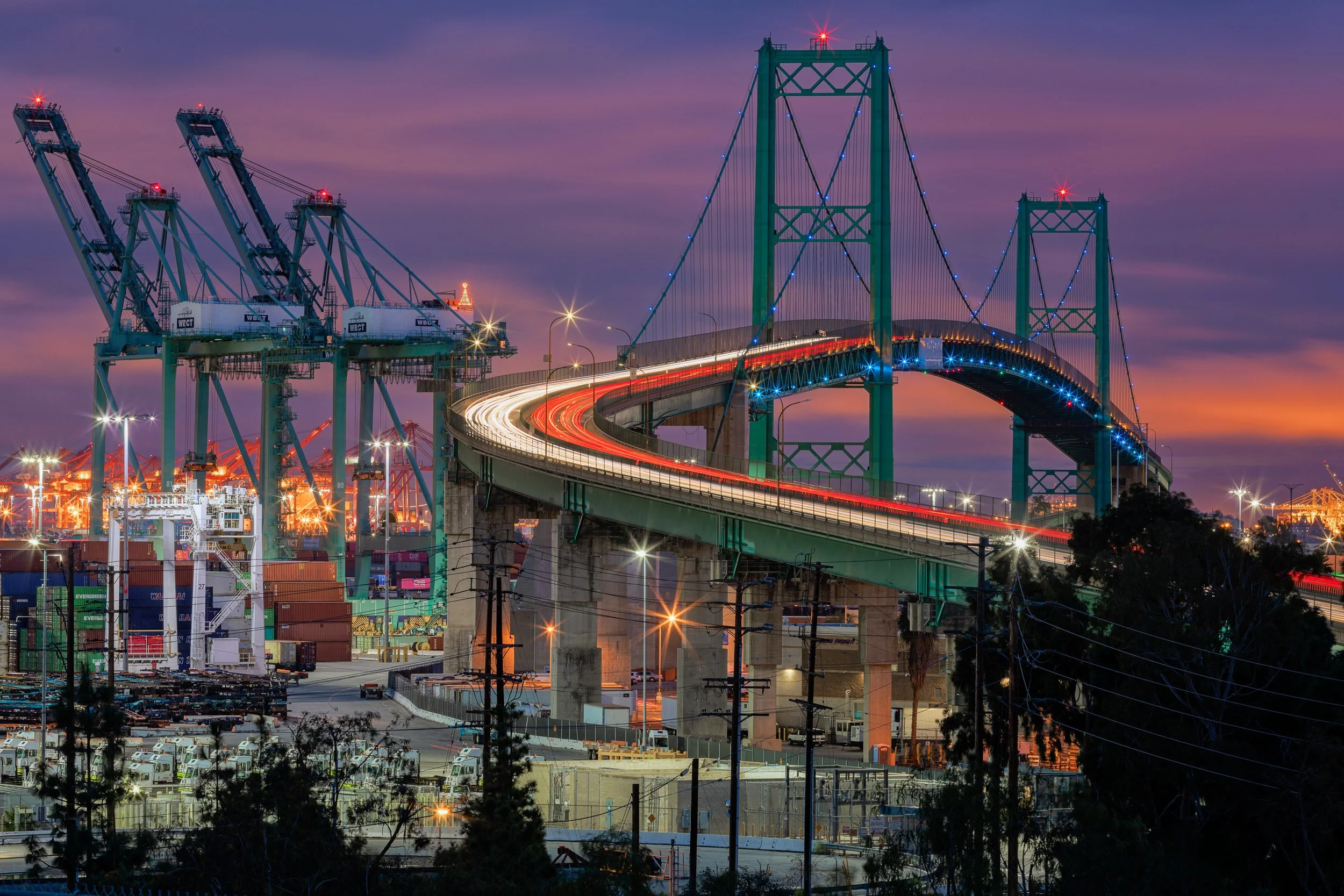 Long exposure light trails curving across the Vincent Thomas Bridge beside illuminated shipping cranes in San Pedro at dusk by Chris Ewen Crosby.
